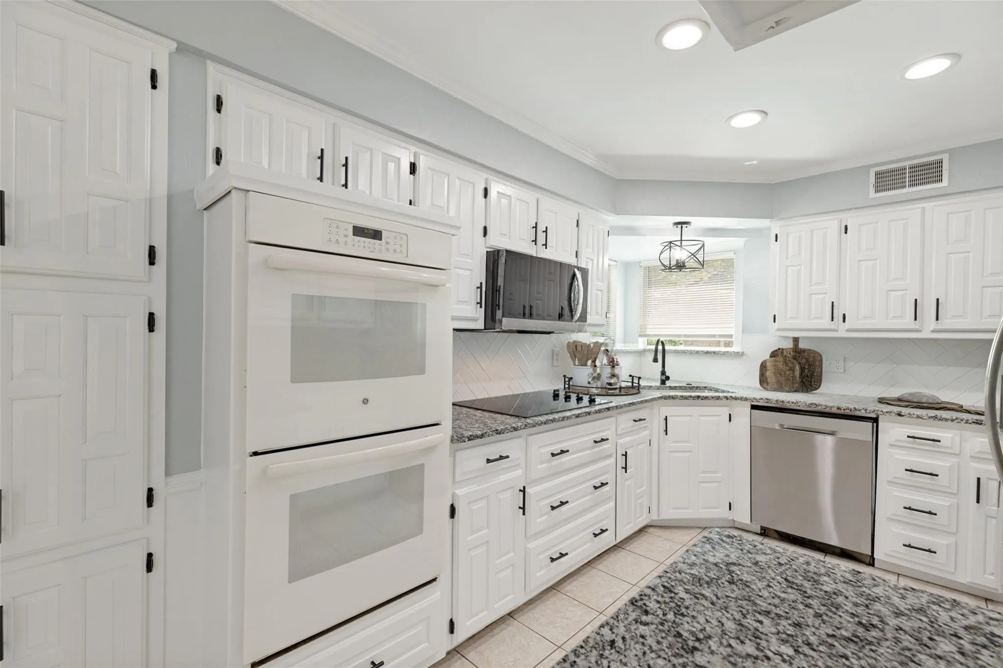 This inviting room features elegant white wainscoting paired with soft neutral walls, creating a refined and timeless look. The rich flooring adds warmth and contrast, while the large windows bring in balanced natural light and offer a pleasant view of the lawn. A stylish metal-frame ceiling fixture adds character overhead, with a coordinating fixture visible in the adjacent space for a cohesive design flow. The doorway leads into a tiled area and a crisp white door, highlighting a smooth transition between rooms and the thoughtful layout of the home. This space stands out for its architectural detailing and bright, welcoming presentation.