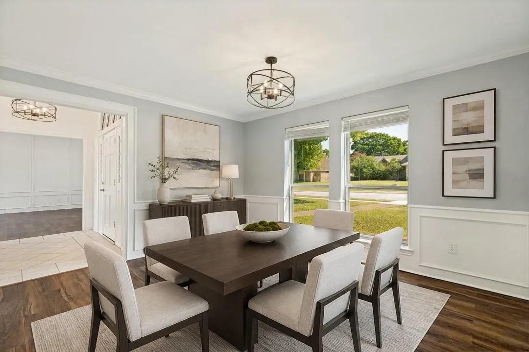This inviting room features elegant white wainscoting paired with soft neutral walls, creating a refined and timeless look. The rich flooring adds warmth and contrast, while the large windows bring in balanced natural light and offer a pleasant view of the lawn. A stylish metal-frame ceiling fixture adds character overhead, with a coordinating fixture visible in the adjacent space for a cohesive design flow. The doorway leads into a tiled area and a crisp white door, highlighting a smooth transition between rooms and the thoughtful layout of the home. This space stands out for its architectural detailing and bright, welcoming presentation.