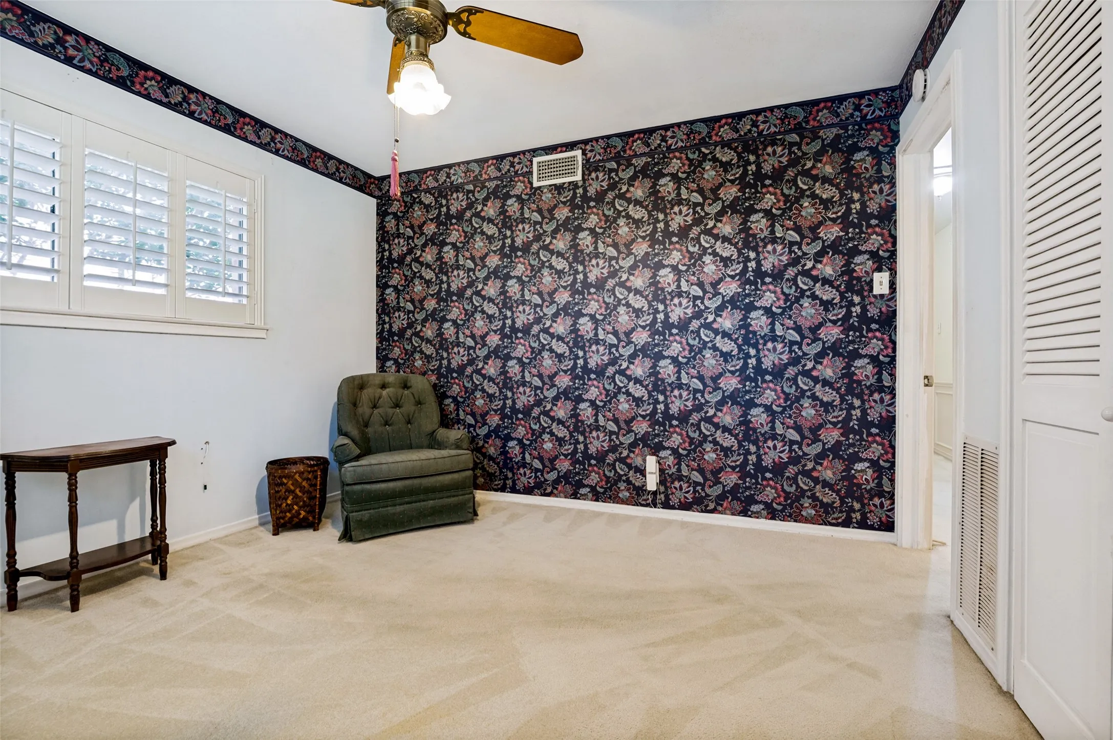 Ceiling fan with light fixture, window with white plantation shutters, patterned accent wall, white louvered doors, and beige carpeting