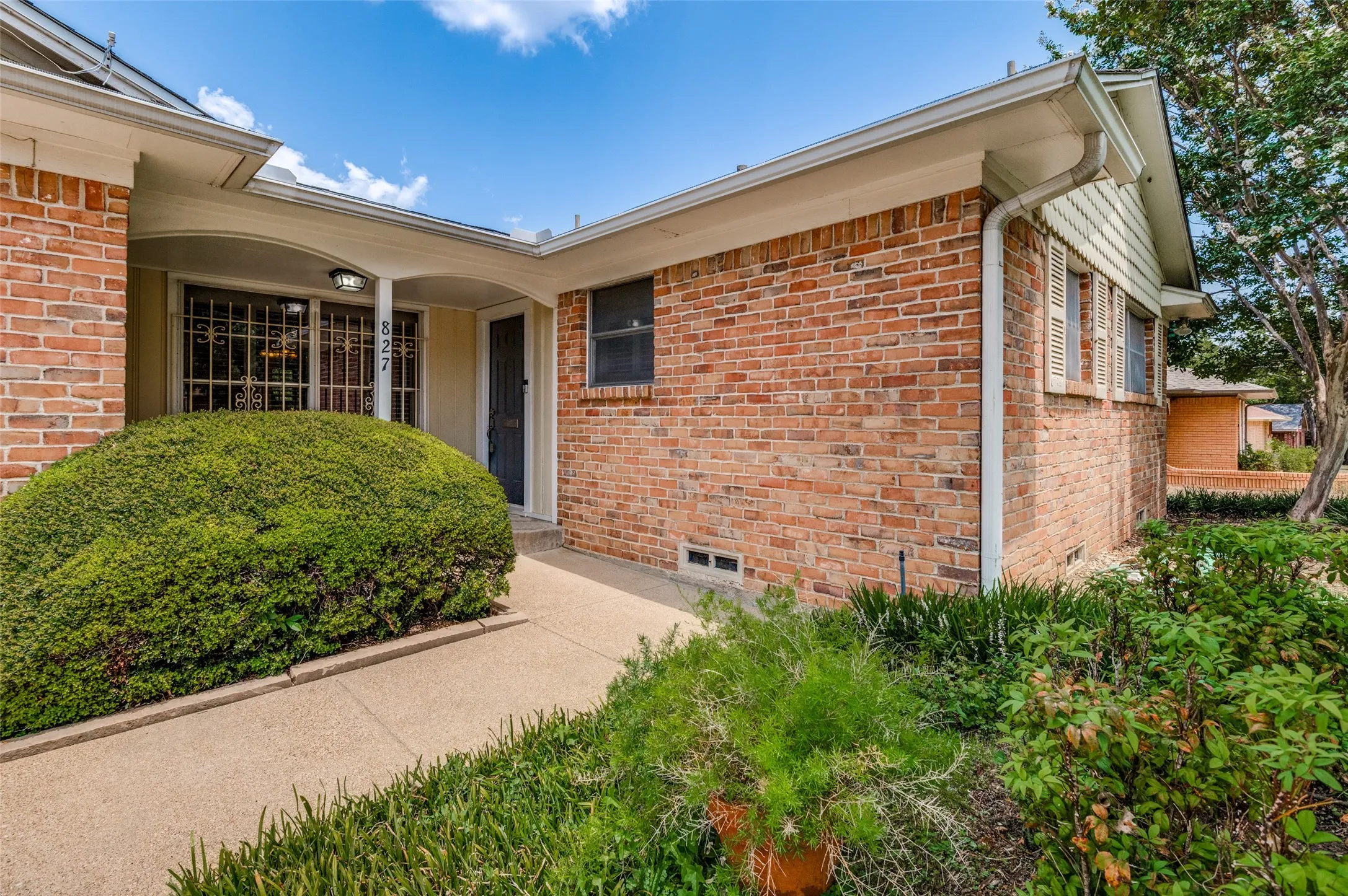 Brick exterior with white trim, covered entry, front door with security screen, and mature landscaping