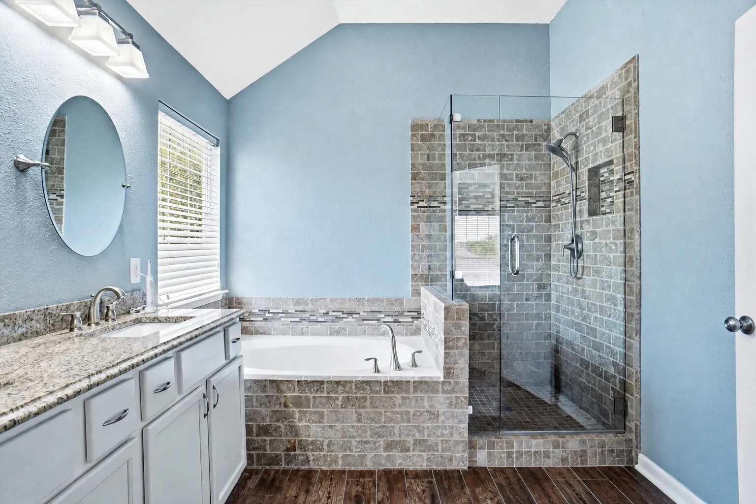 Primary bathroom featuring granite countertops, dual vanities, a soaking tub, and a separate glass-enclosed walk-in shower with travertine tile.