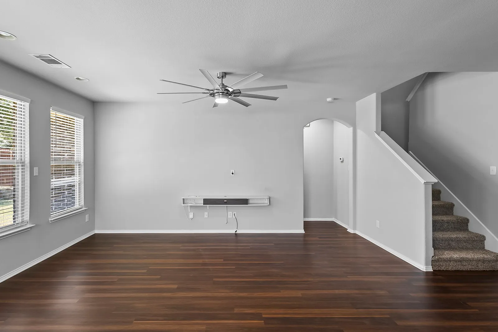 Spacious living area featuring wood-finish flooring, a contemporary ceiling fan, two windows with blinds, a built-in media console, and a carpeted staircase