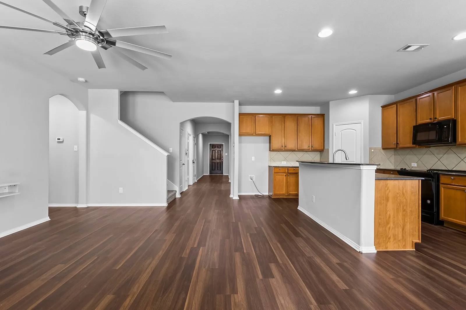 Open-concept living area featuring wood-finish flooring, a contemporary ceiling fan, and a kitchen with wood cabinetry, a breakfast bar, and dark​​‌​​​​‌​​‌‌​‌‌​​​‌‌​‌​‌​‌​​​‌​​ countertops