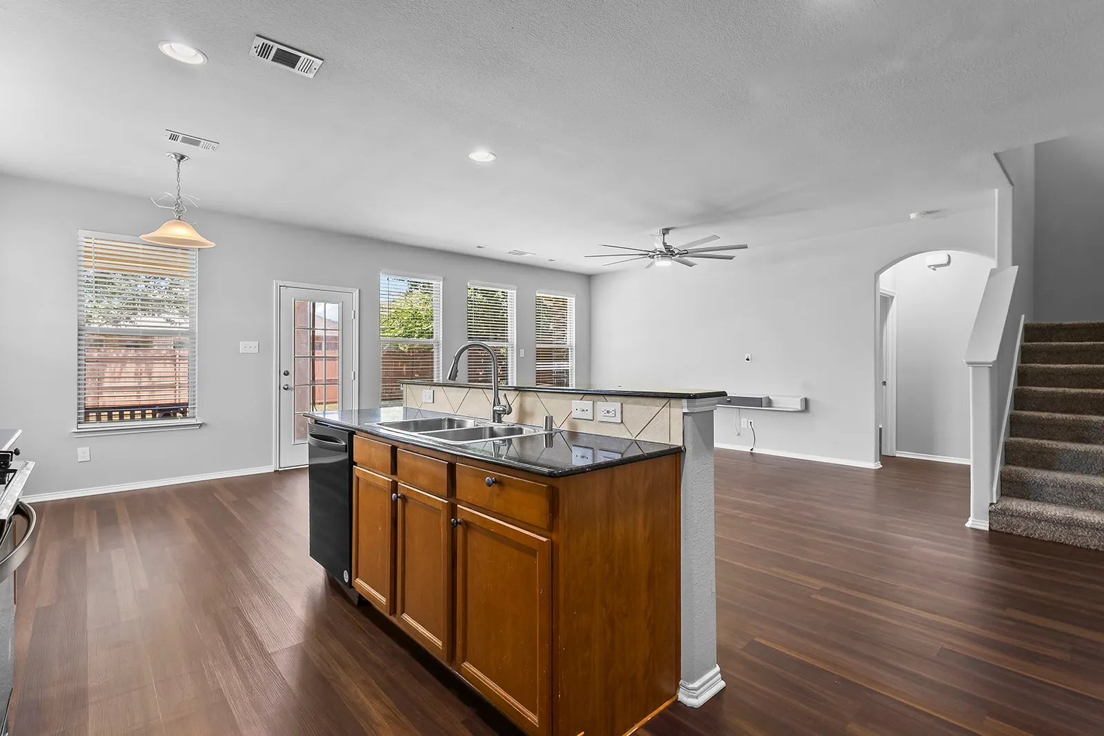 Open-concept living space featuring wood-finish flooring, a kitchen island with integrated sink, black dishwasher, and a tiled backsplash