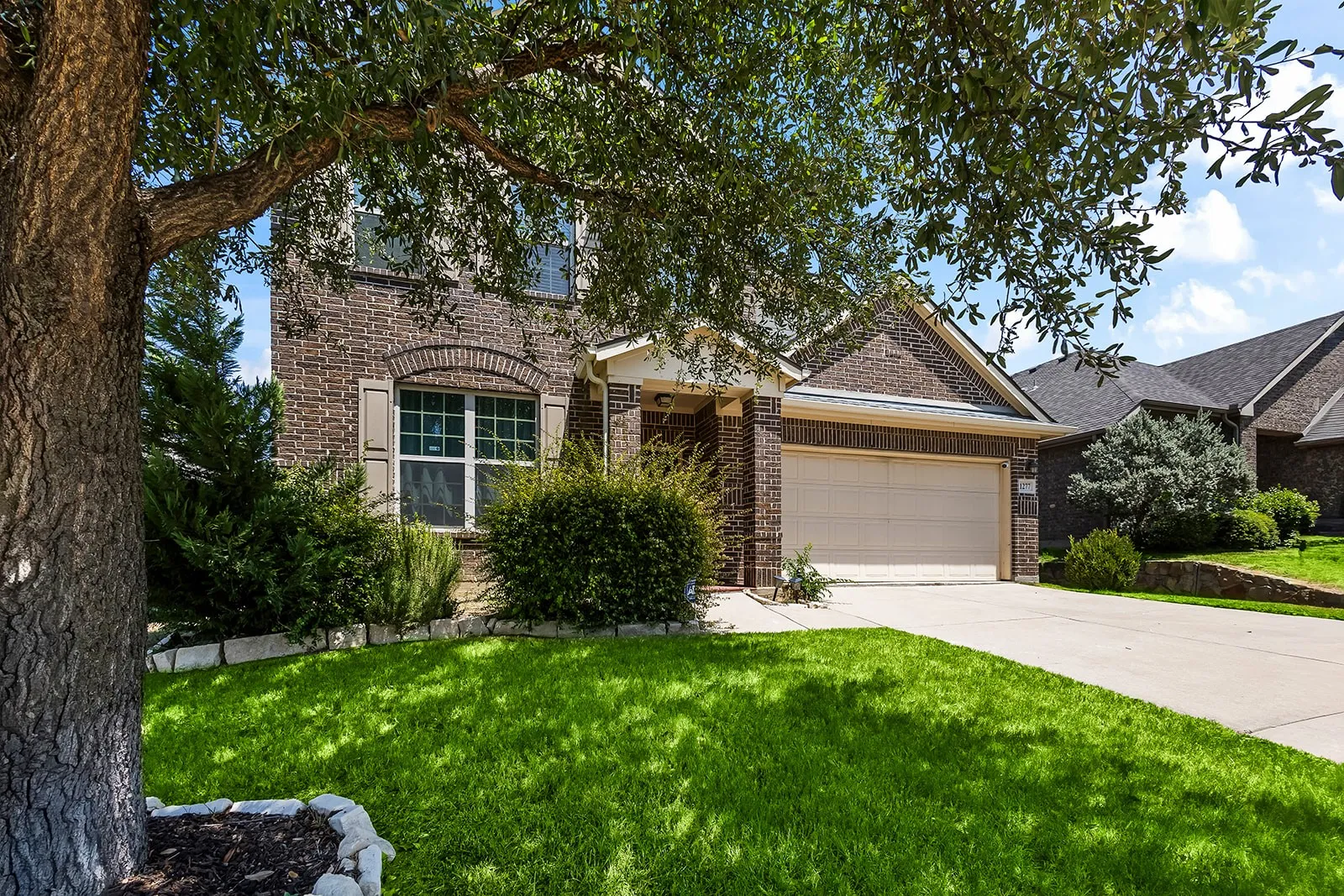 Brick-clad exterior featuring a two-car garage, arched window accents, mature trees, manicured lawn, and a concrete driveway