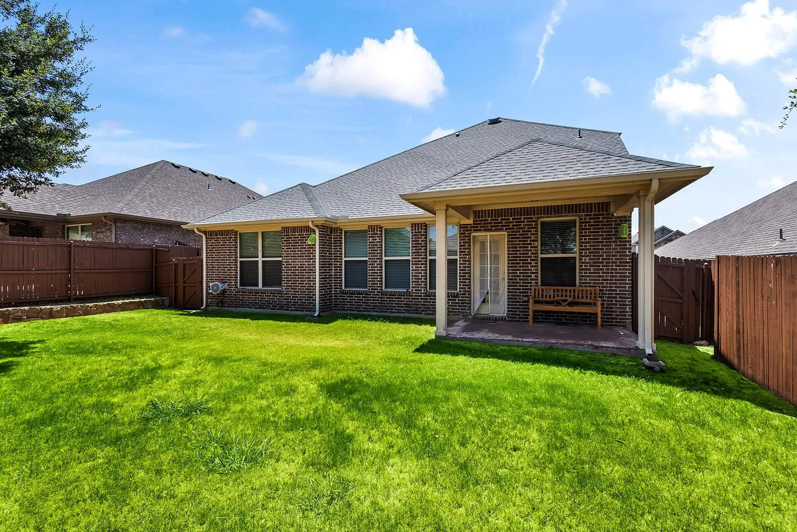 Spacious rear yard featuring a covered patio, brick exterior, multiple windows, dark wood fencing, and a manicured lawn