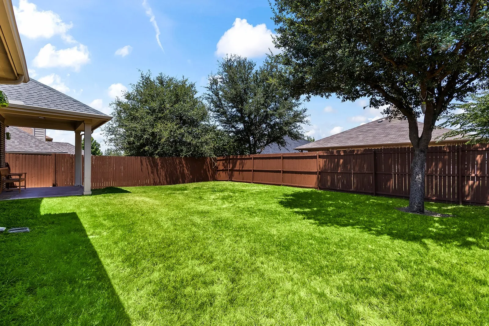 Expansive backyard featuring a lush green lawn, mature trees, a covered patio with concrete flooring, and a privacy fence