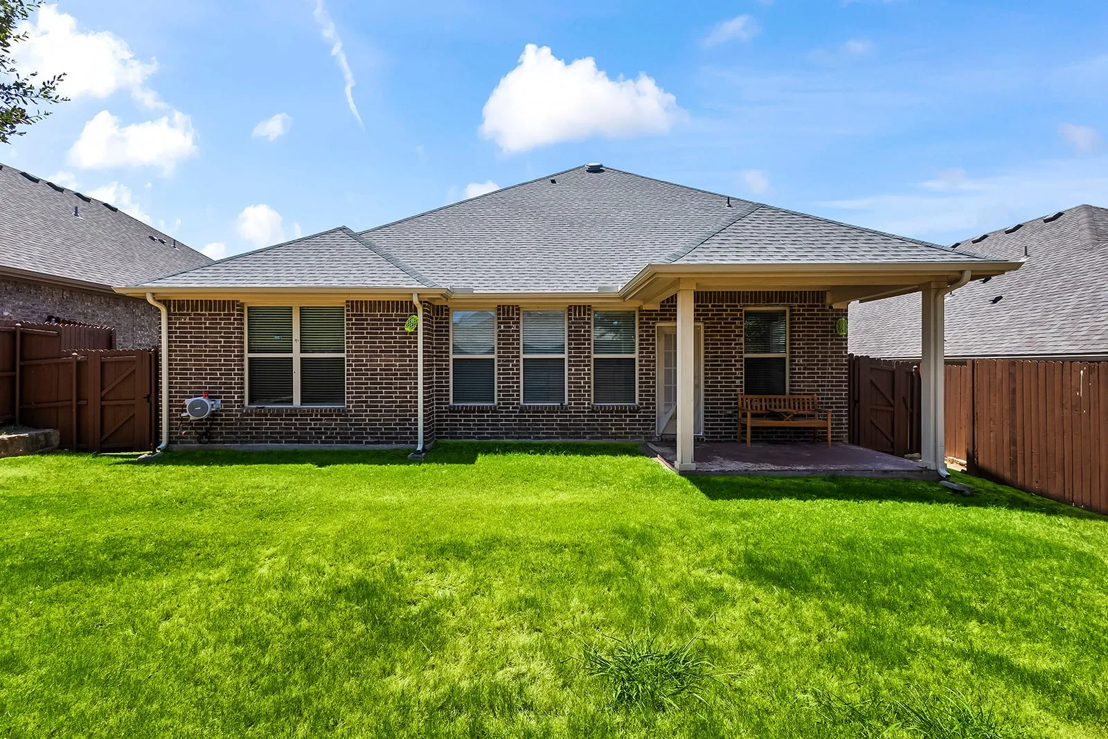 Manicured green lawn with a covered patio, brick exterior, multiple windows, and a wood fence