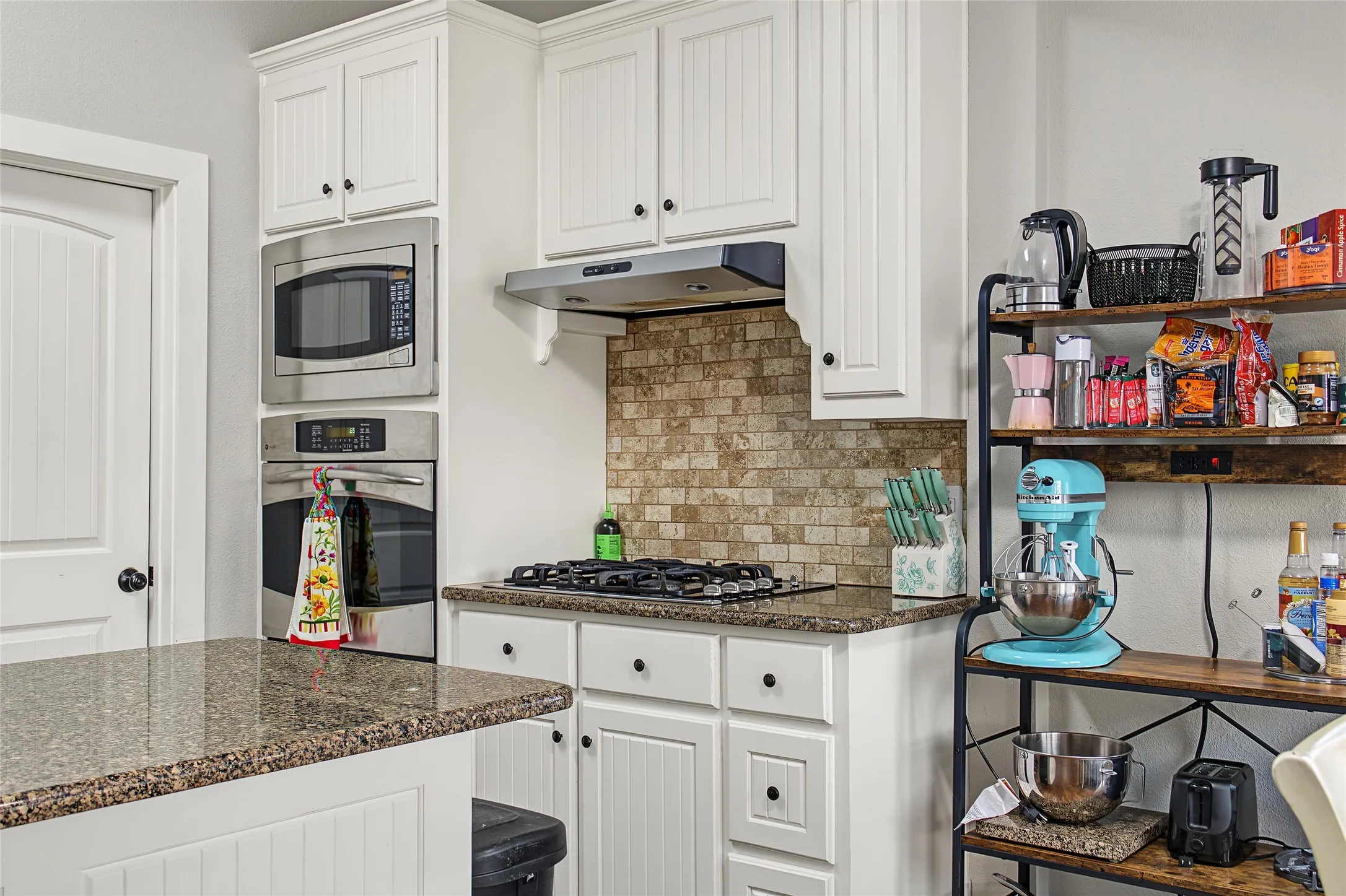Built-in stainless steel microwave and oven, gas cooktop, tile backsplash, white cabinetry with beadboard detailing, and granite countertops