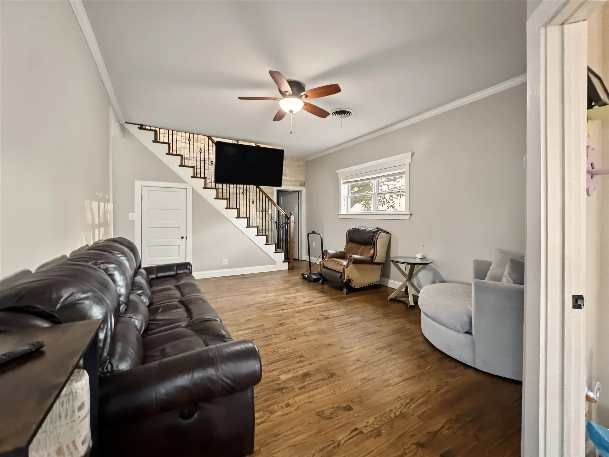 Wood-finish flooring, staircase with metal balusters, ceiling fan, crown molding, and a window with decorative trim