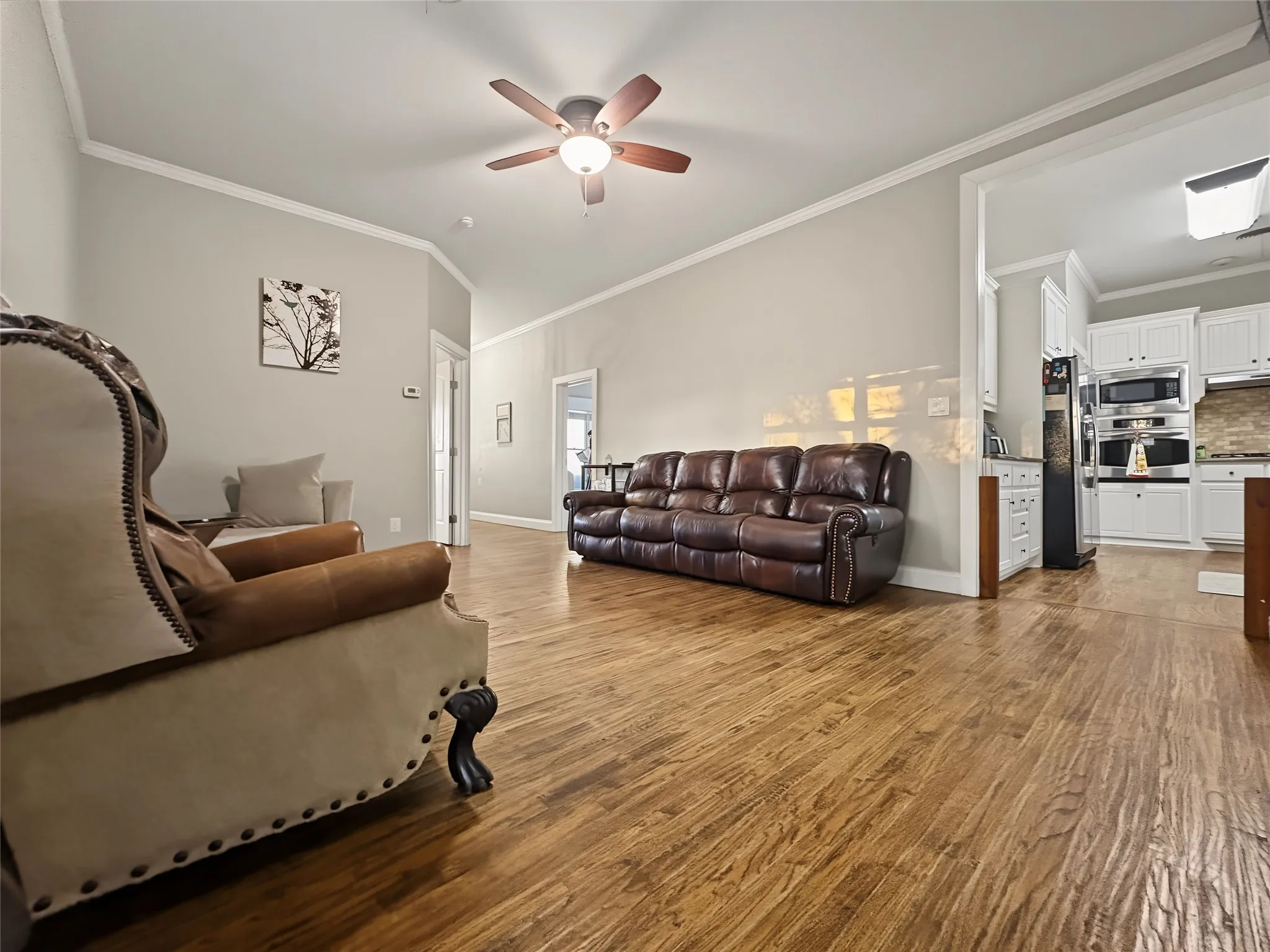 Wood-finish flooring throughout, vaulted ceiling with a ceiling fan, crown molding, and an open concept layout with an adjacent kitchen featuring white cabinetry and built-in appliances