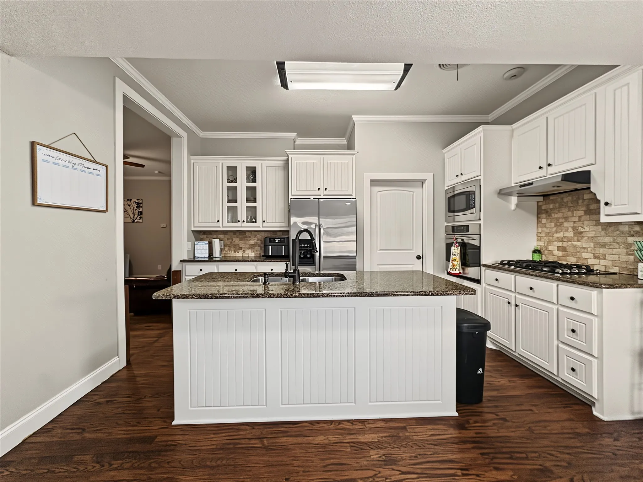 Kitchen featuring a central island with a sink, white cabinetry, a tiled backsplash, built-in stainless steel appliances, and wood-finish flooring