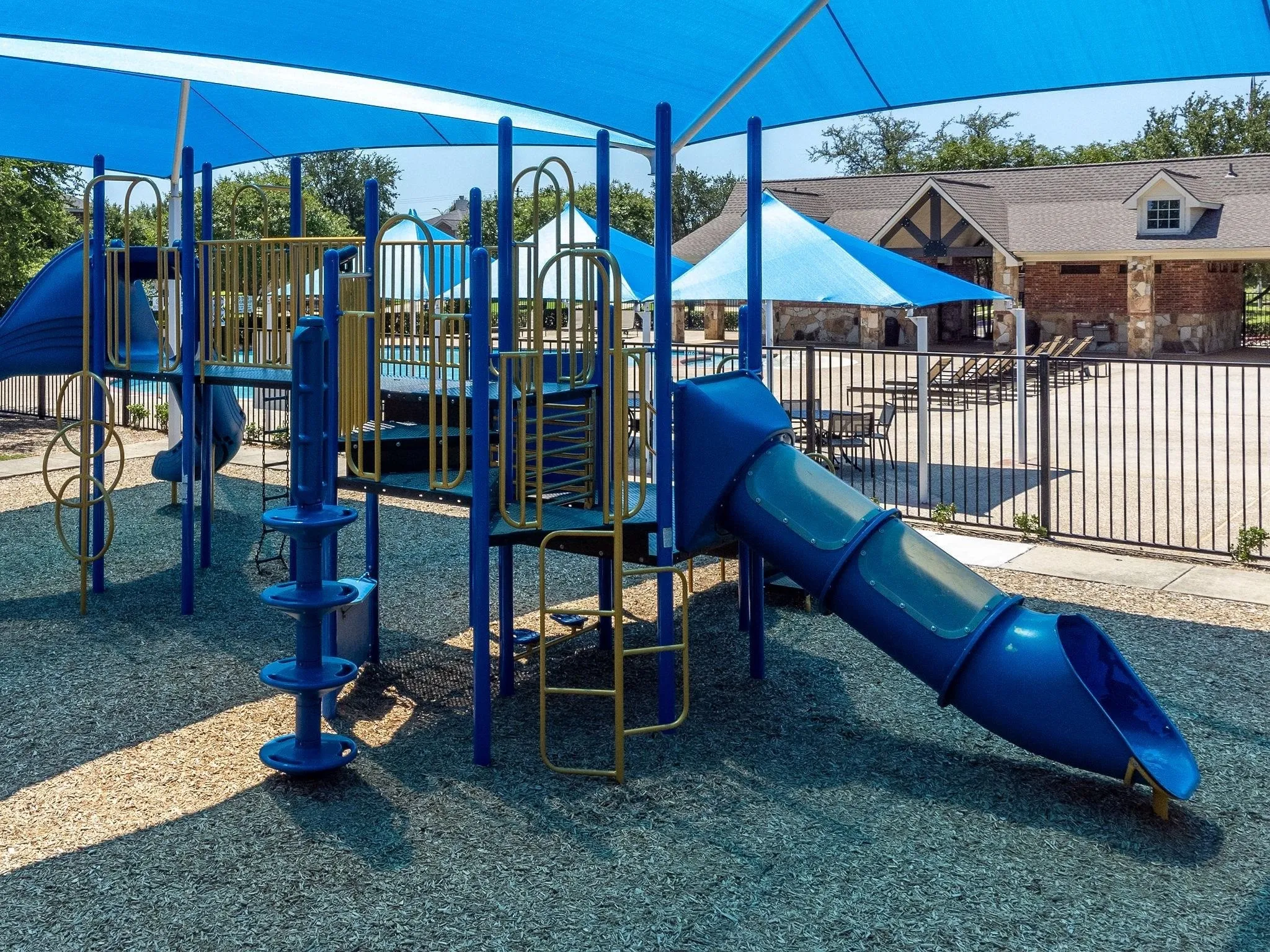 Playground featuring a multi-level play structure with slides and climbing elements, covered by a blue shade canopy, and surrounded by protective ground surfacing