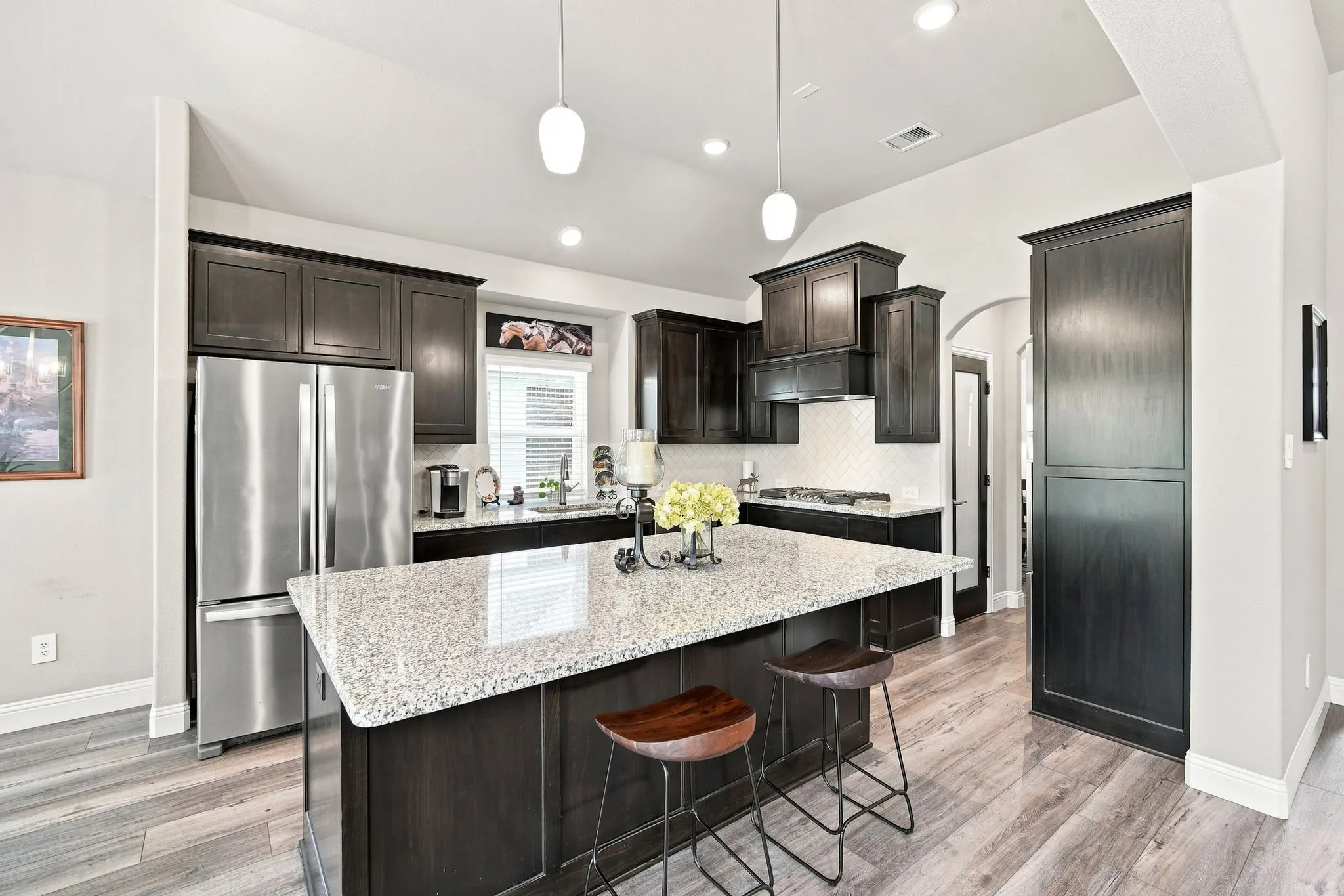 The kitchen featuring dark wood cabinetry, light-toned granite countertops, stainless steel appliances, a tile backsplash, and wood-finish flooring
