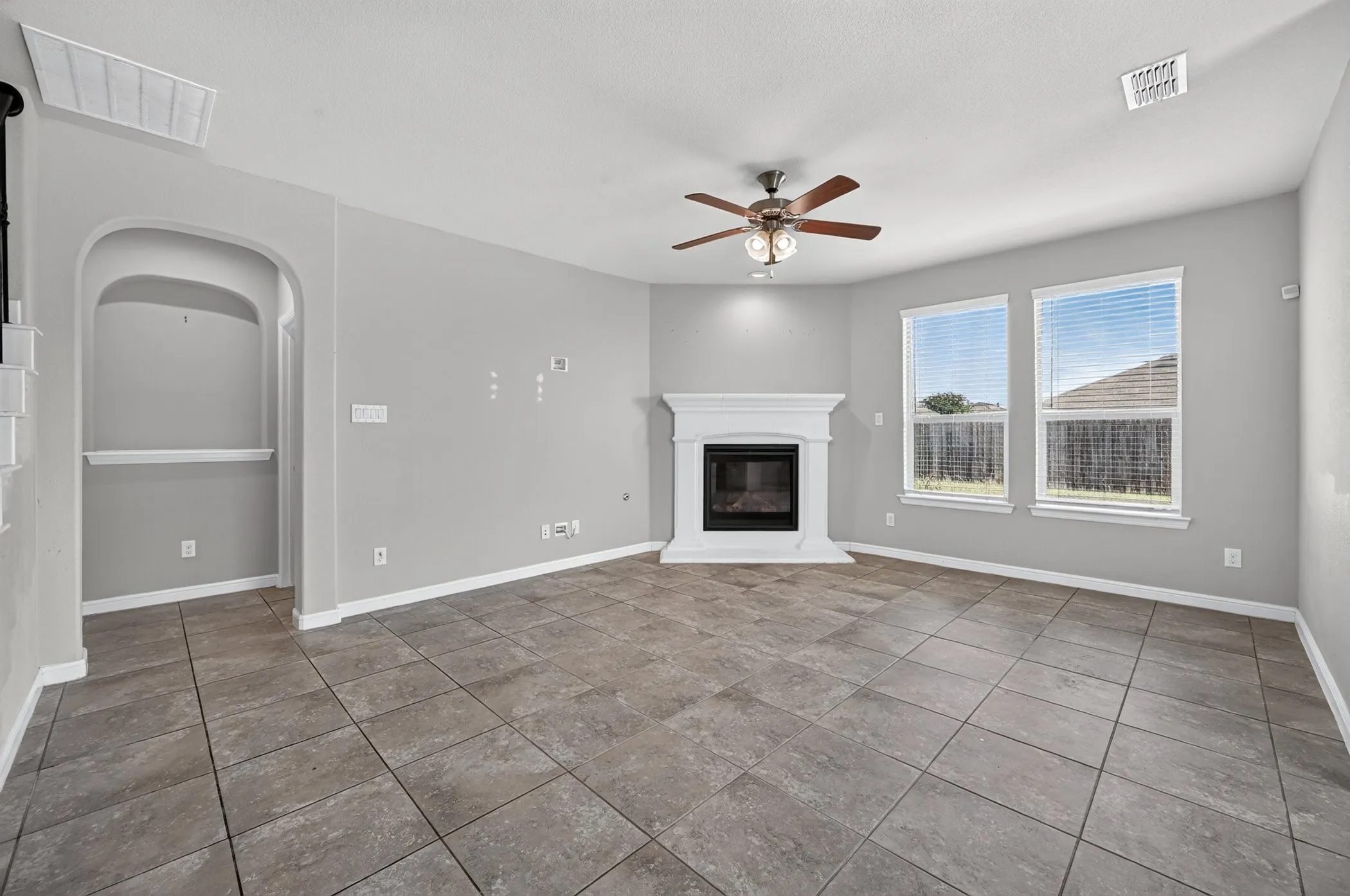 Spacious living area showcasing diagonal tile flooring, a ceiling fan with integrated lighting, large dual windows with natural light, a corner fireplace with white surround and dark insert, and an arched entryway with architectural detail