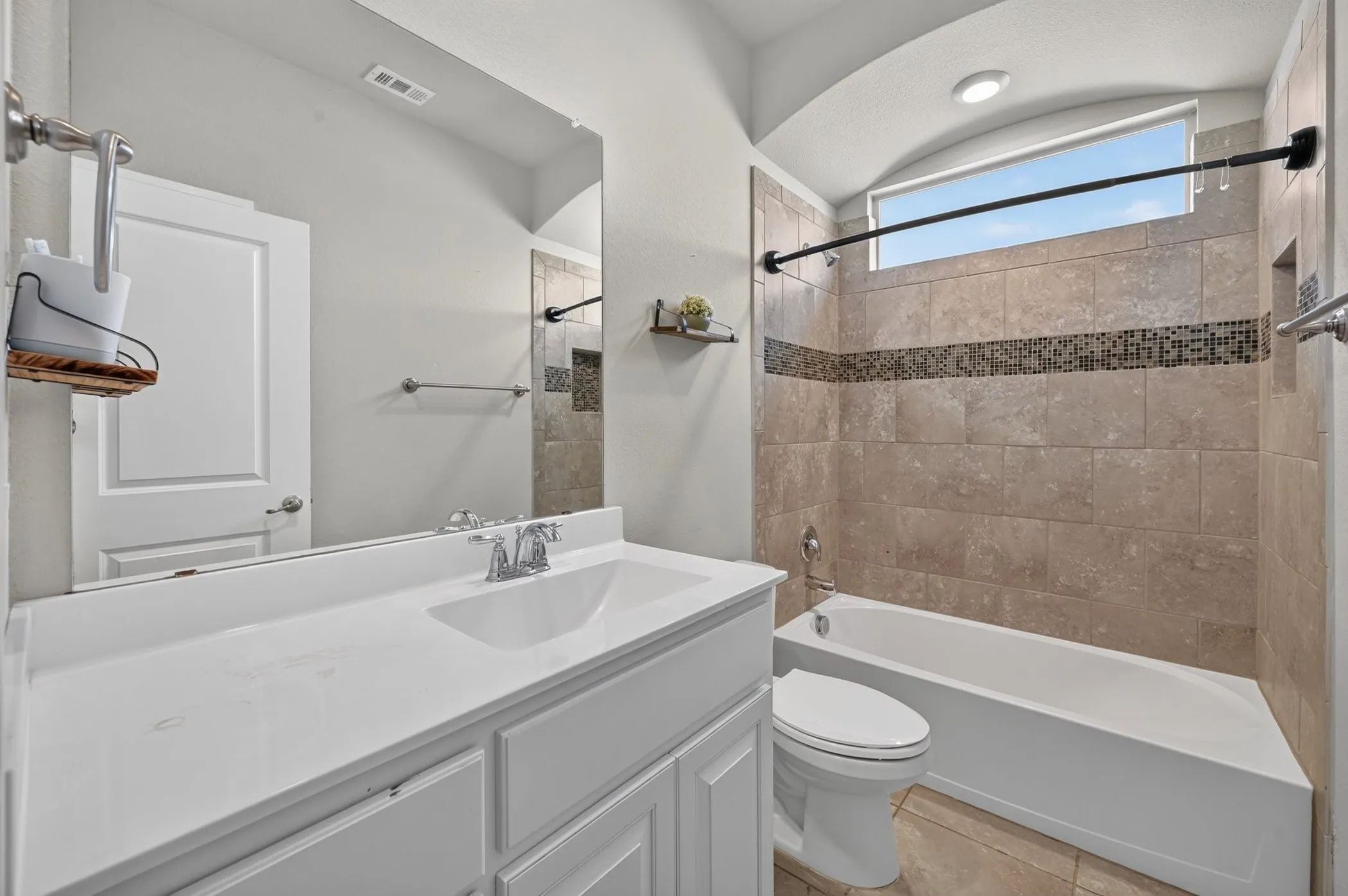 White vanity with integrated sink, chrome faucet, rectangular mirror, and a tiled shower-tub combination with a decorative mosaic band