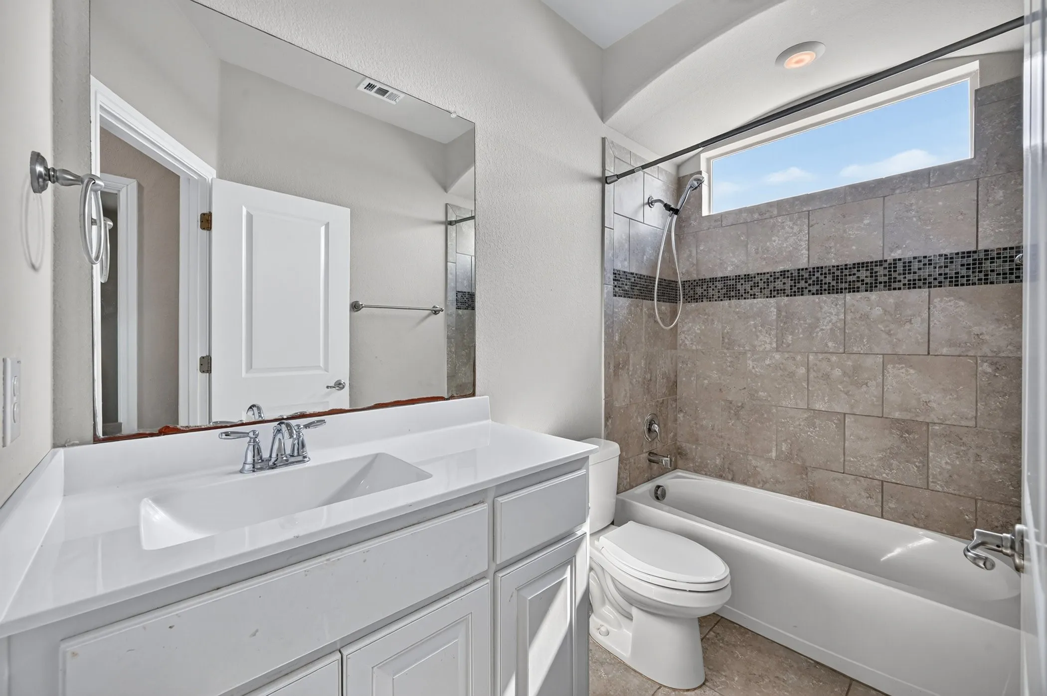 Built-in vanity with integrated sink, expansive mirror, bathtub with tiled shower surround, decorative tile accent band, and an arched ceiling alcove