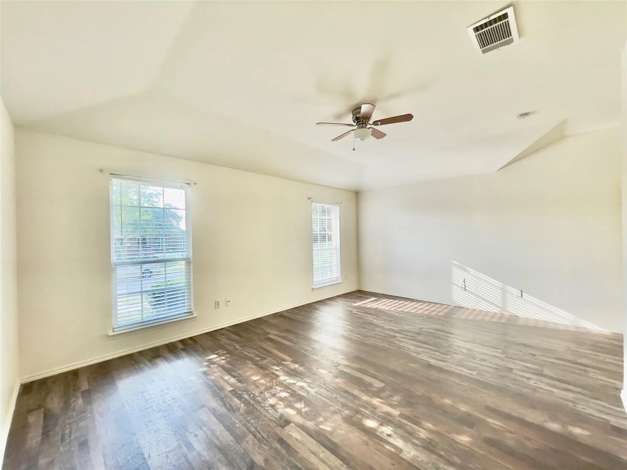 Bright and spacious room featuring hardwood flooring, abundant natural light from dual windows, ceiling fan with integrated lighting, and clean, neutral walls throughout