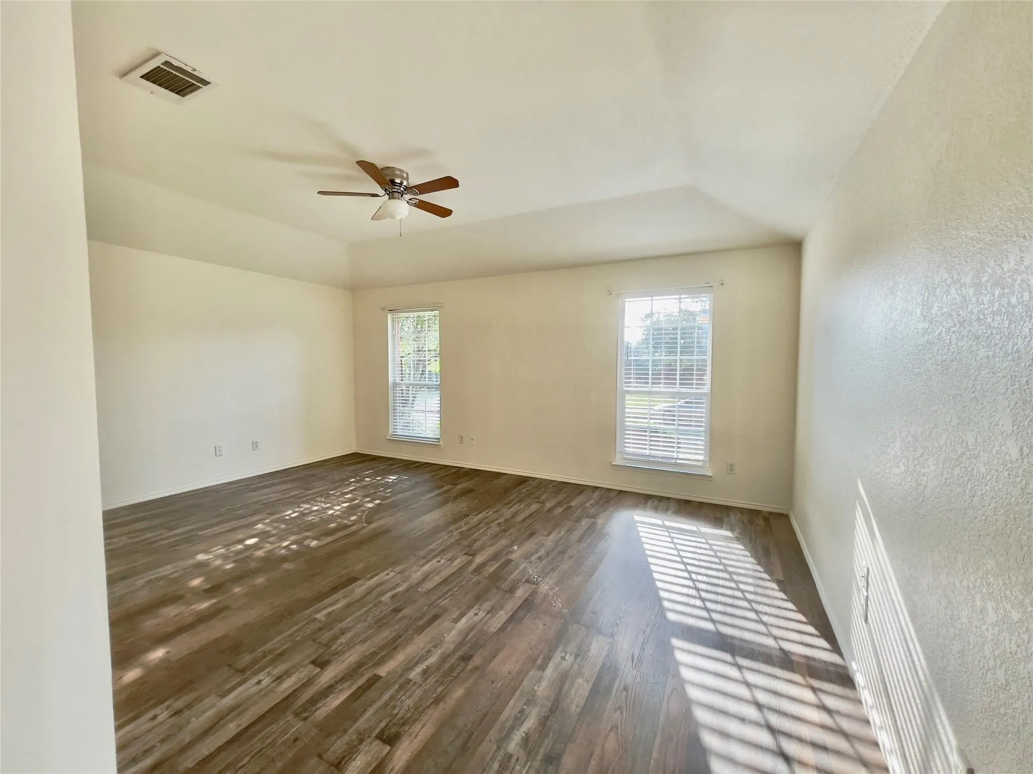 Wood-finish flooring throughout, two large windows with blinds, ceiling fan with integrated lighting, and a vaulted ceiling