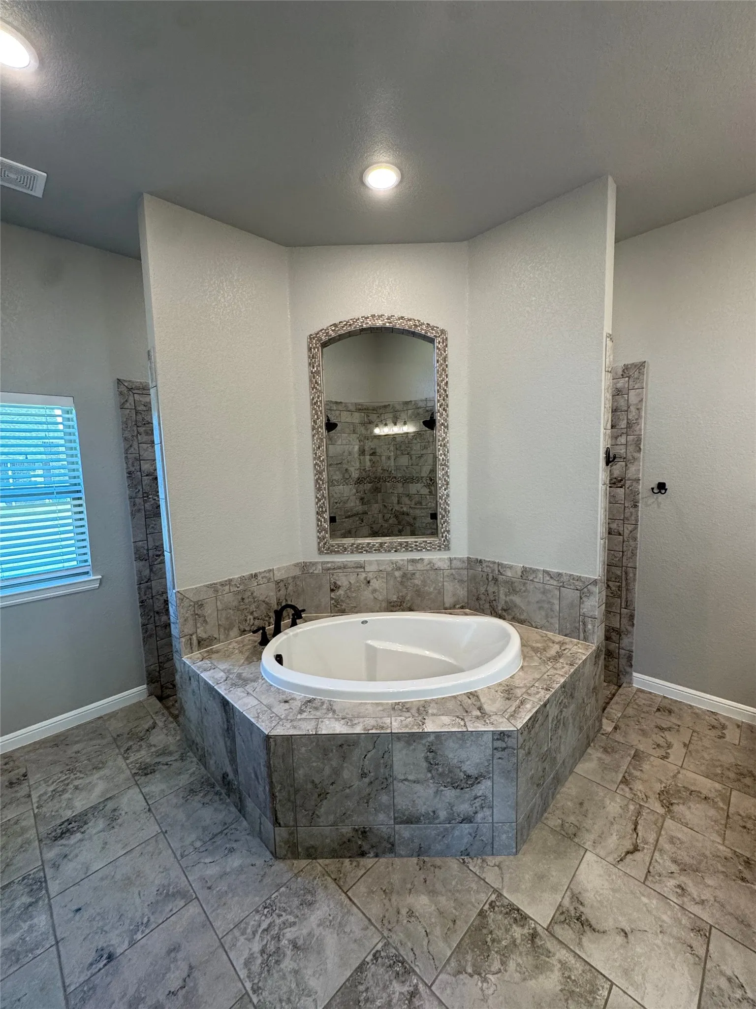 Recessed lighting, tiled flooring, built-in soaking tub, tiled tub surround, and an arched mirror with a decorative frame