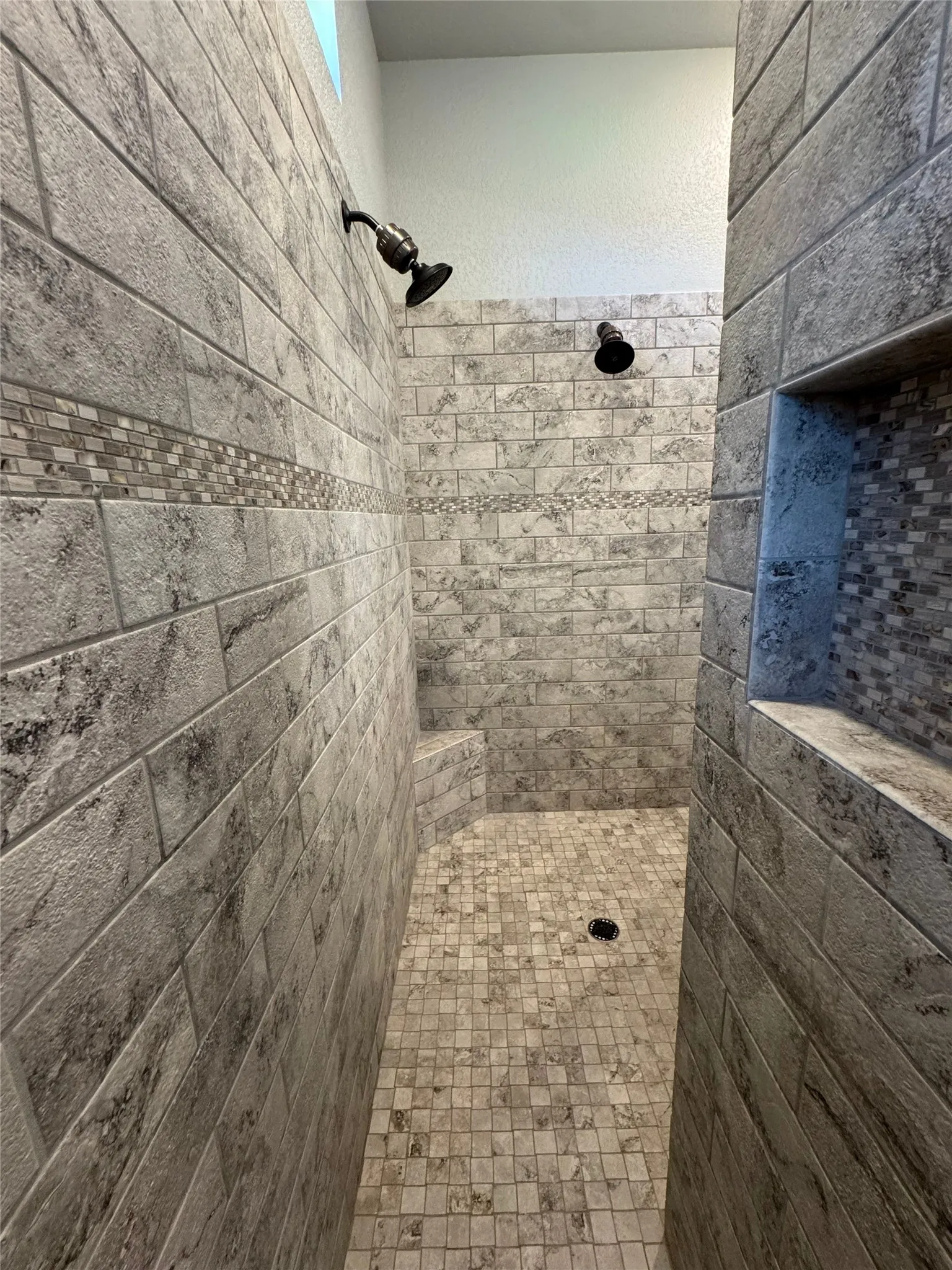 Tiled shower enclosure featuring dual shower heads, a built-in bench, a recessed niche, and mosaic tile accents