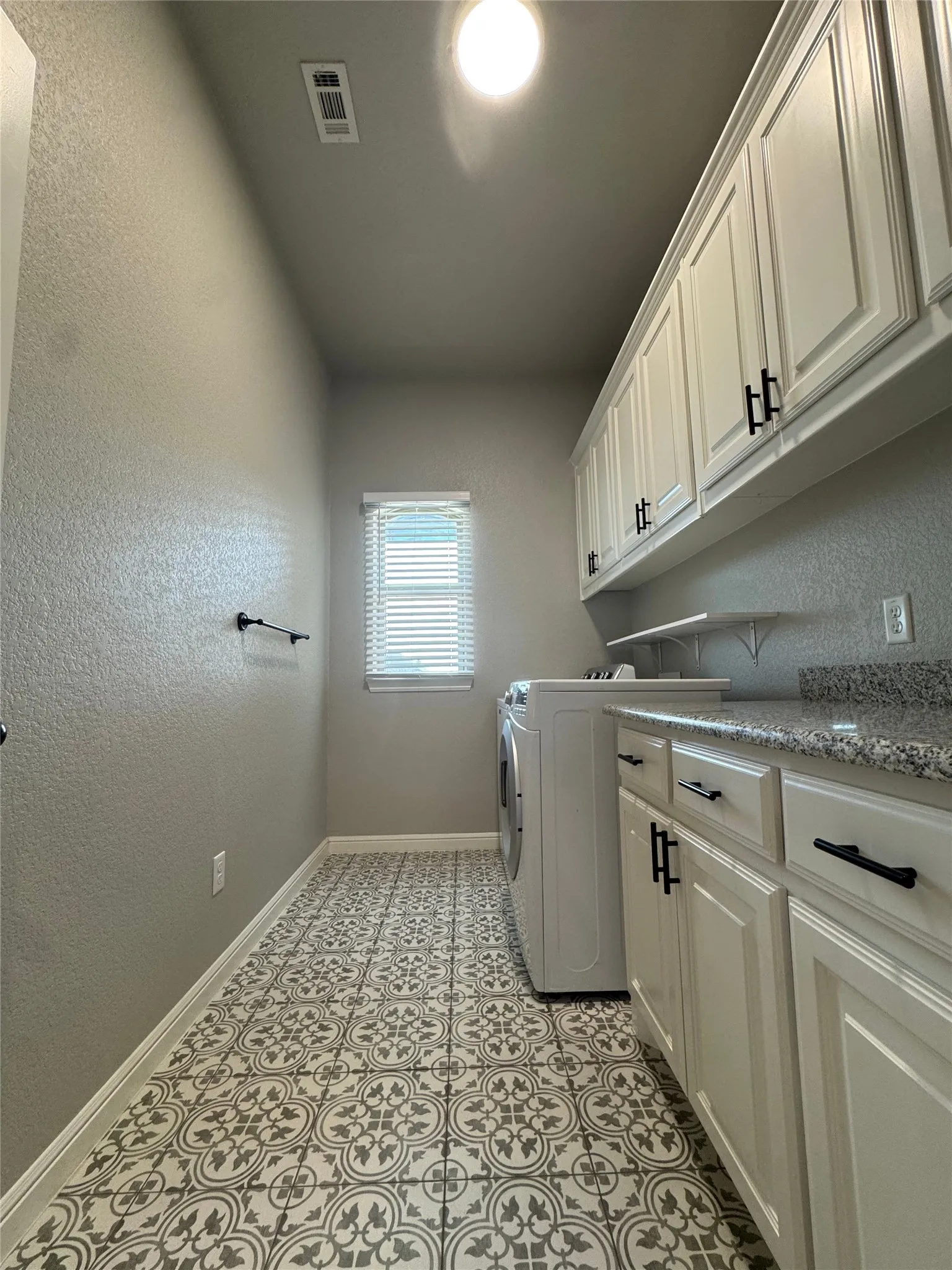 Patterned tile flooring, shaker-style cabinetry with matte black hardware, granite-style countertop, and a single window with blinds