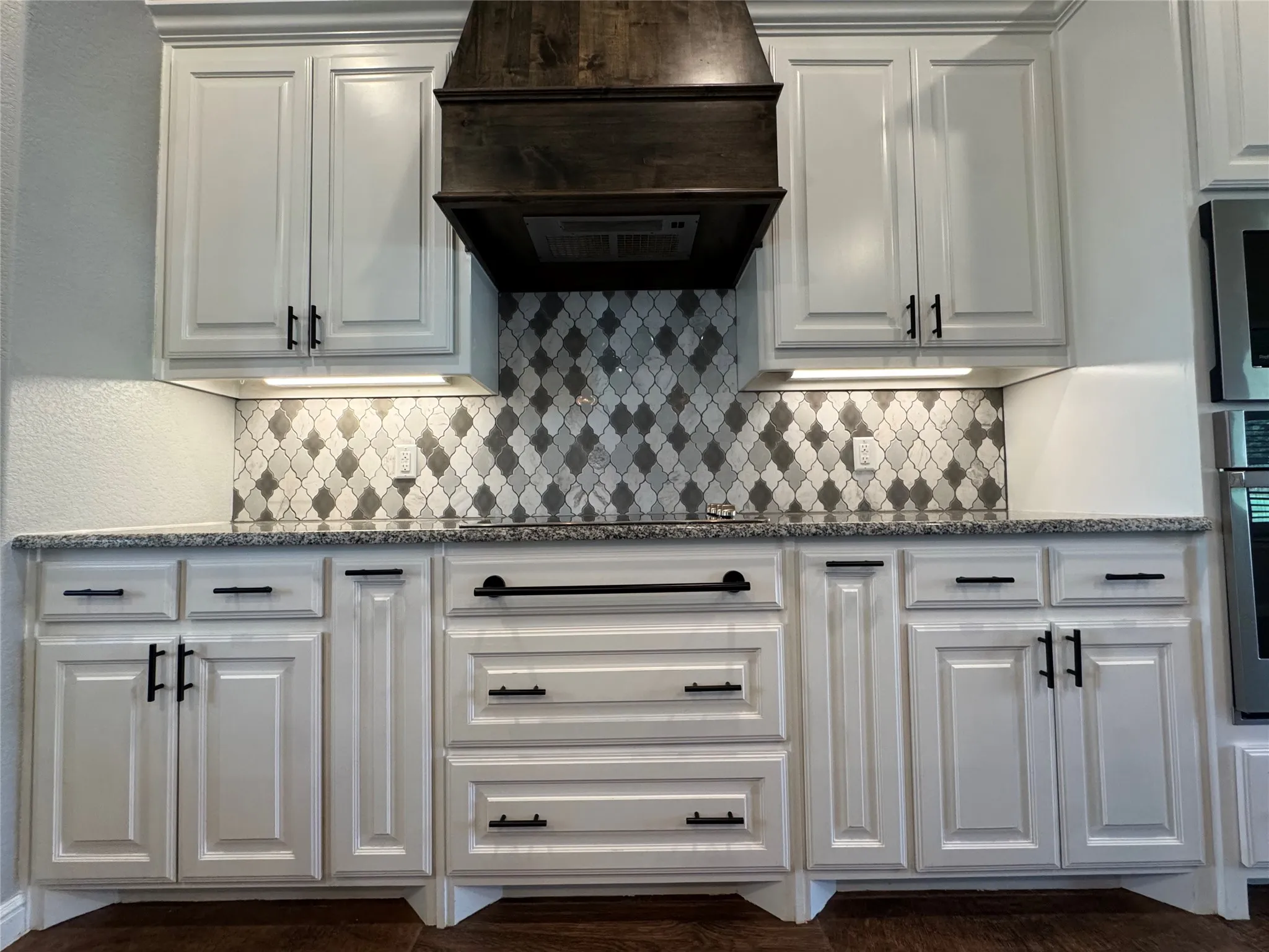 White cabinetry with matte black hardware, mosaic tile backsplash, wood-finish range hood, and speckled countertops