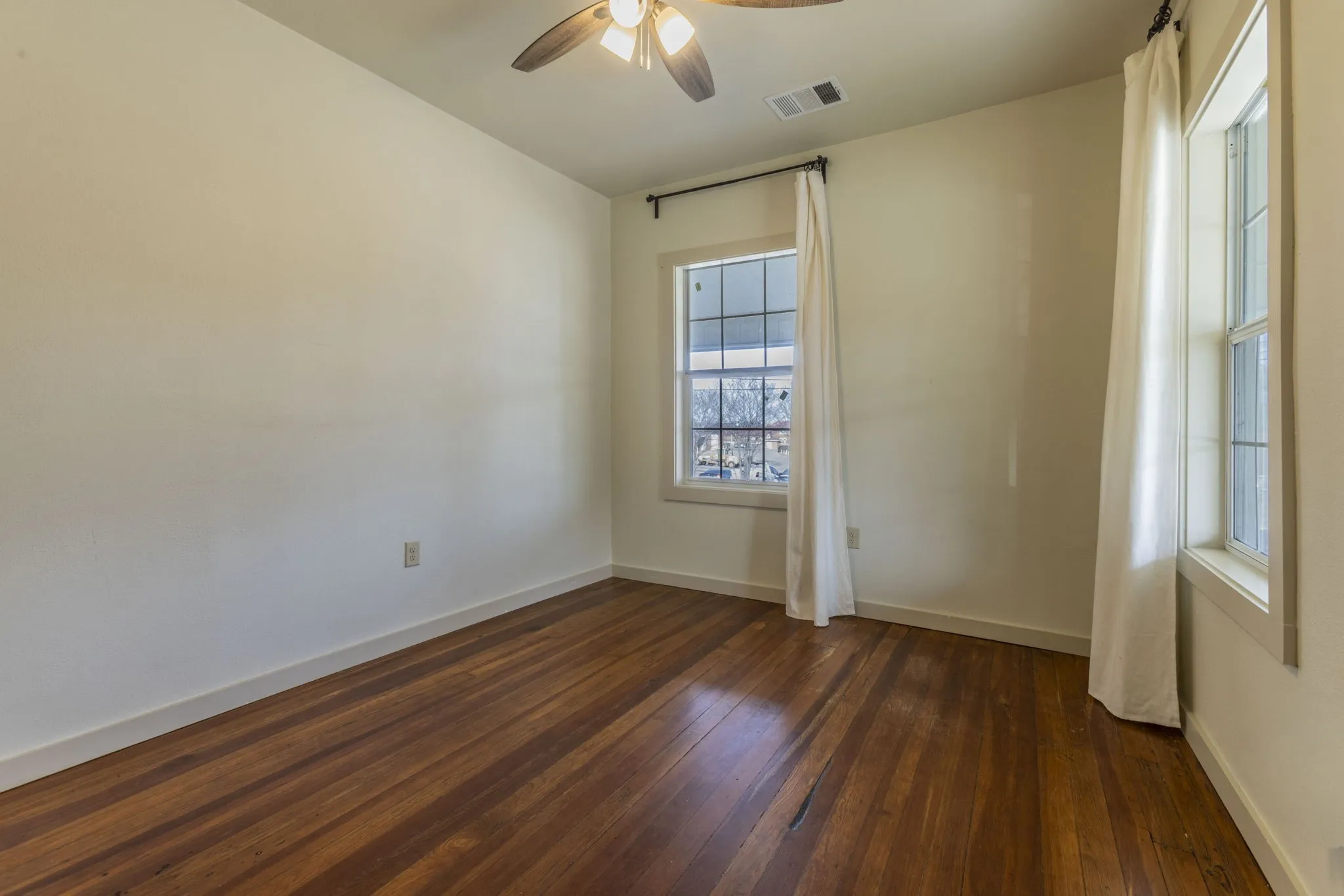 Room featuring rich wood-finish flooring, two window openings, a ceiling fan with integrated lighting, and painted walls with white baseboards