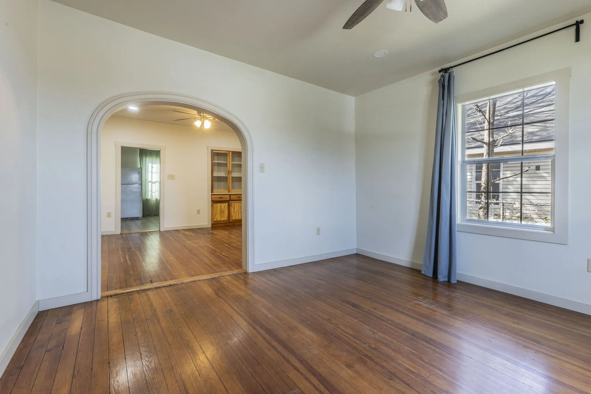 Hardwood flooring throughout, featuring an arched doorway, white walls, a ceiling fan, and a window with white trim