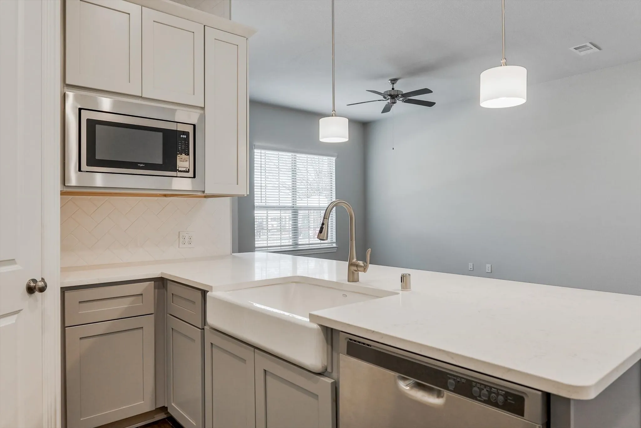 White farmhouse sink, stainless steel microwave, stainless steel dishwasher, white subway tile backsplash, and light-toned countertops