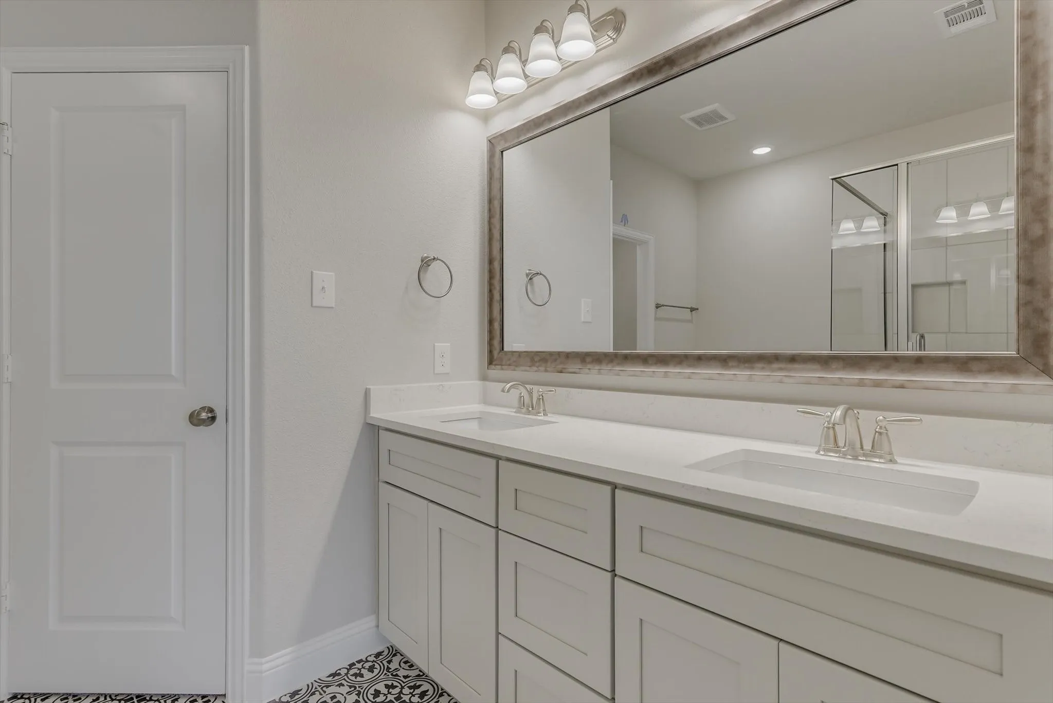 Dual vanity with white cabinetry and integrated sinks, brushed nickel fixtures, a large framed mirror, and patterned floor tiles