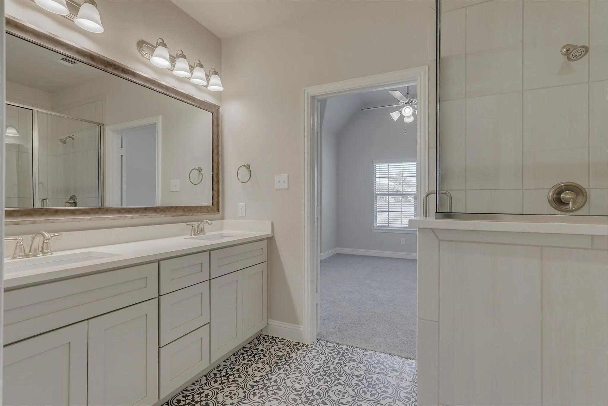 Dual vanity with stone countertops, decorative tile flooring, framed mirror with vanity lighting, and a glass-enclosed shower