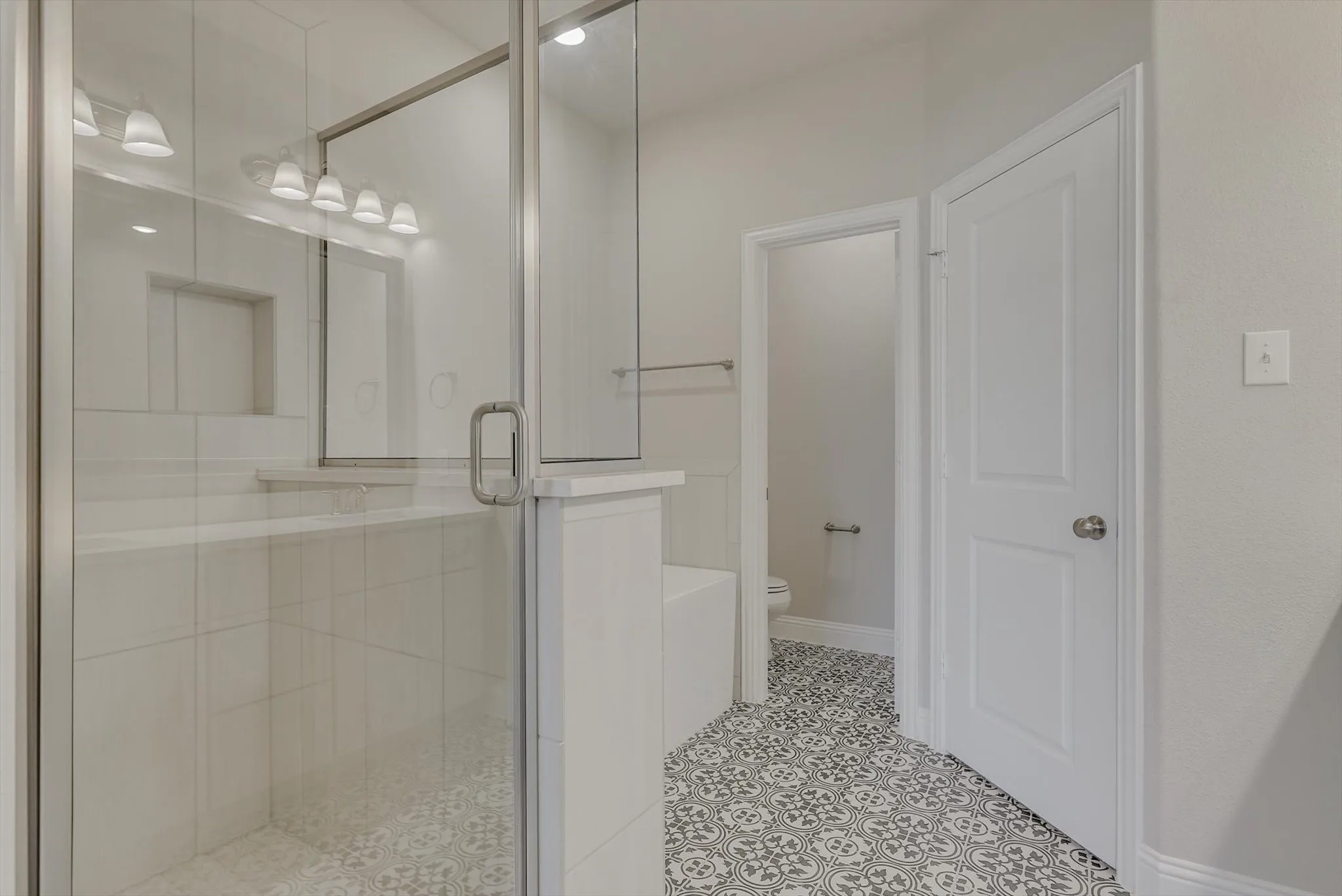 Glass-enclosed shower with white tiled walls and built-in shelving, complemented by patterned floor tiling