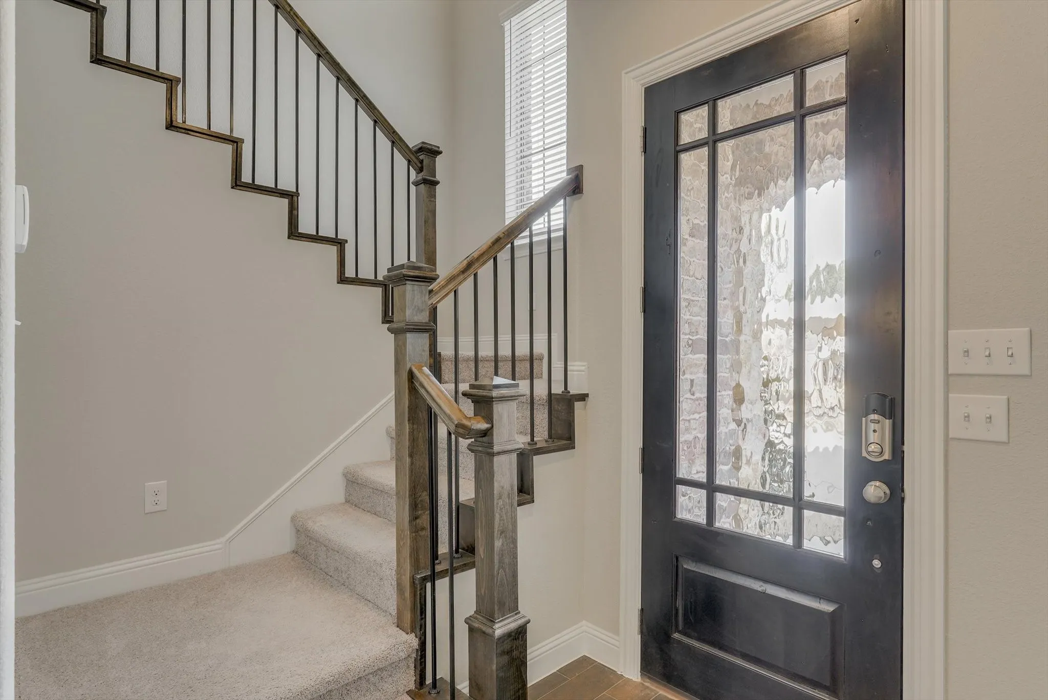 Entry featuring a dark-paneled door with patterned glass, wood-finish flooring, and a carpeted staircase with wood-finish handrails and black metal balusters