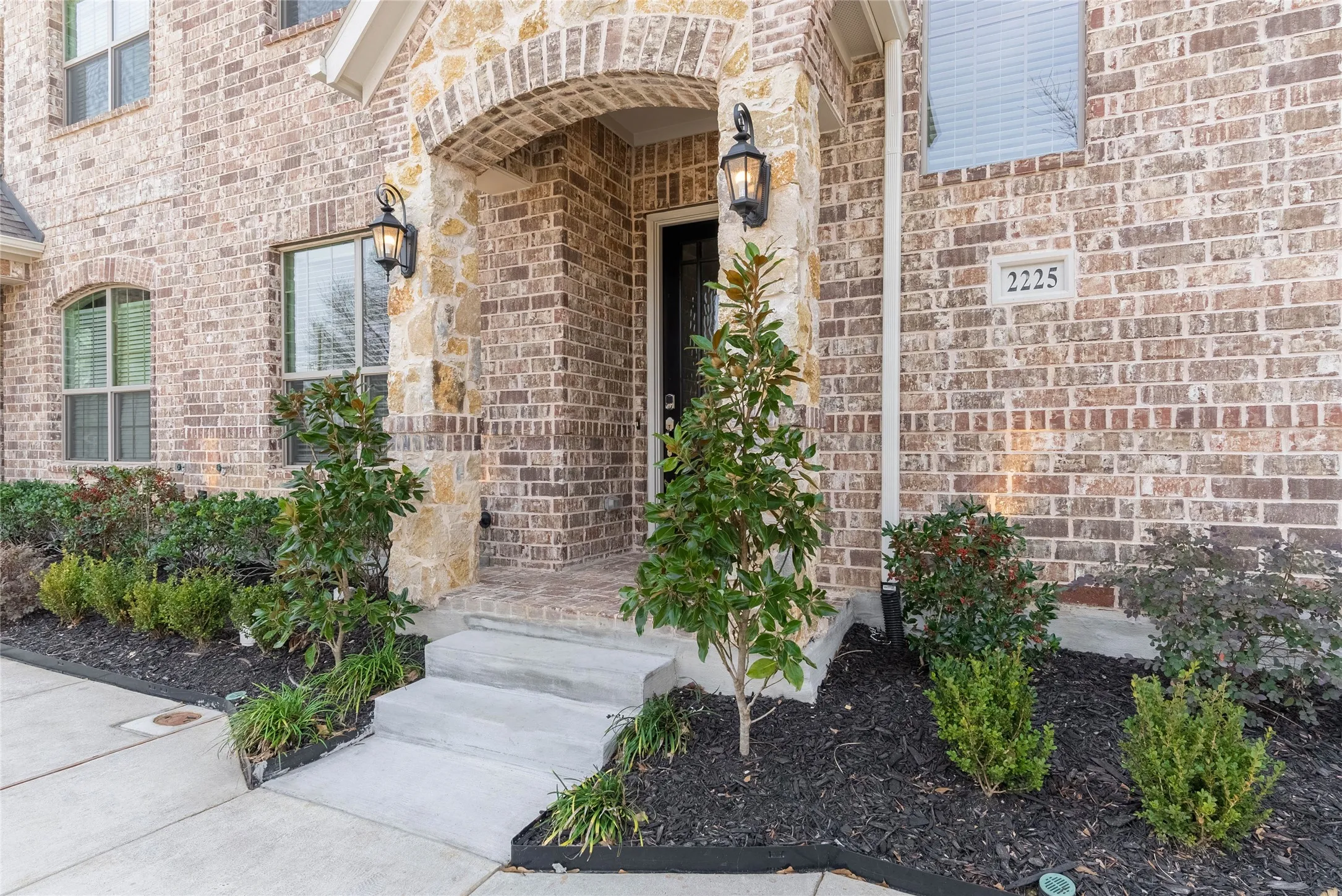 Brick and stone exterior with arched entryway, concrete steps, and landscaped planting beds