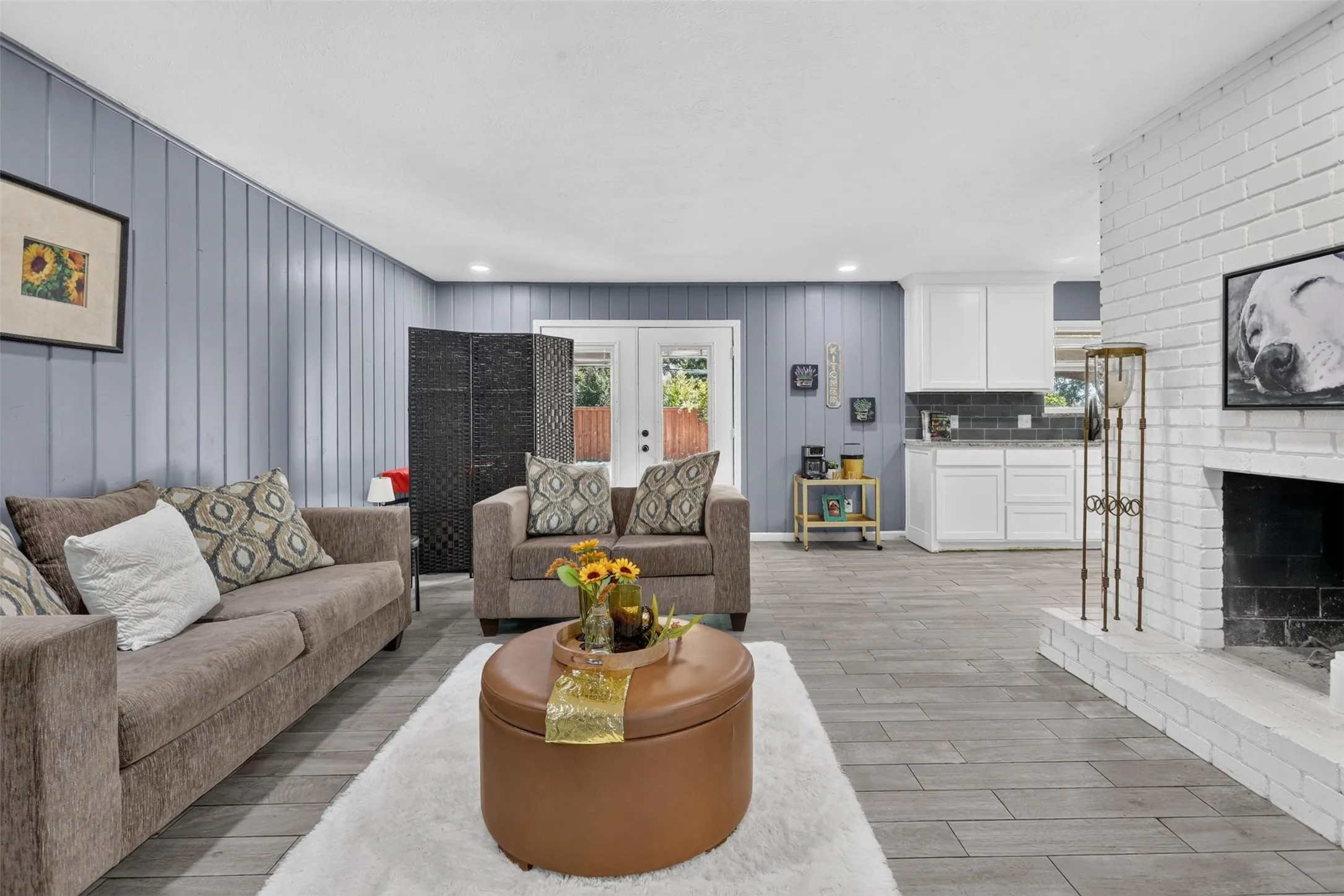 Open-concept living space featuring gray wood-finish flooring throughout, neutral upholstered seating, a round cognac-leather ottoman, and a fireplace with white shiplap surround