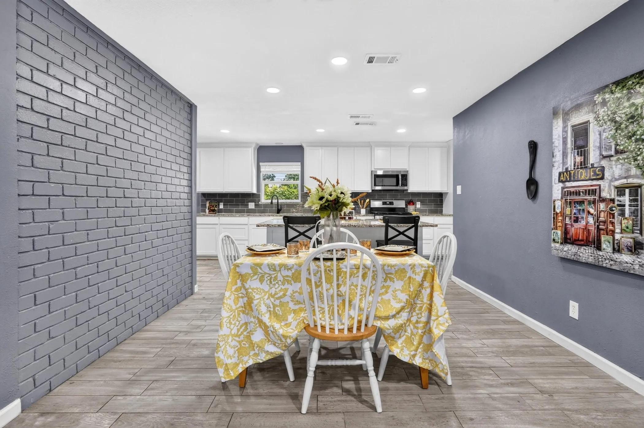 Open-concept dining and kitchen area featuring recessed ceiling lighting, gray painted accent walls, white cabinetry with stainless steel appliances, wood-finish flooring, and a brick accent wall with modern geometric detailing