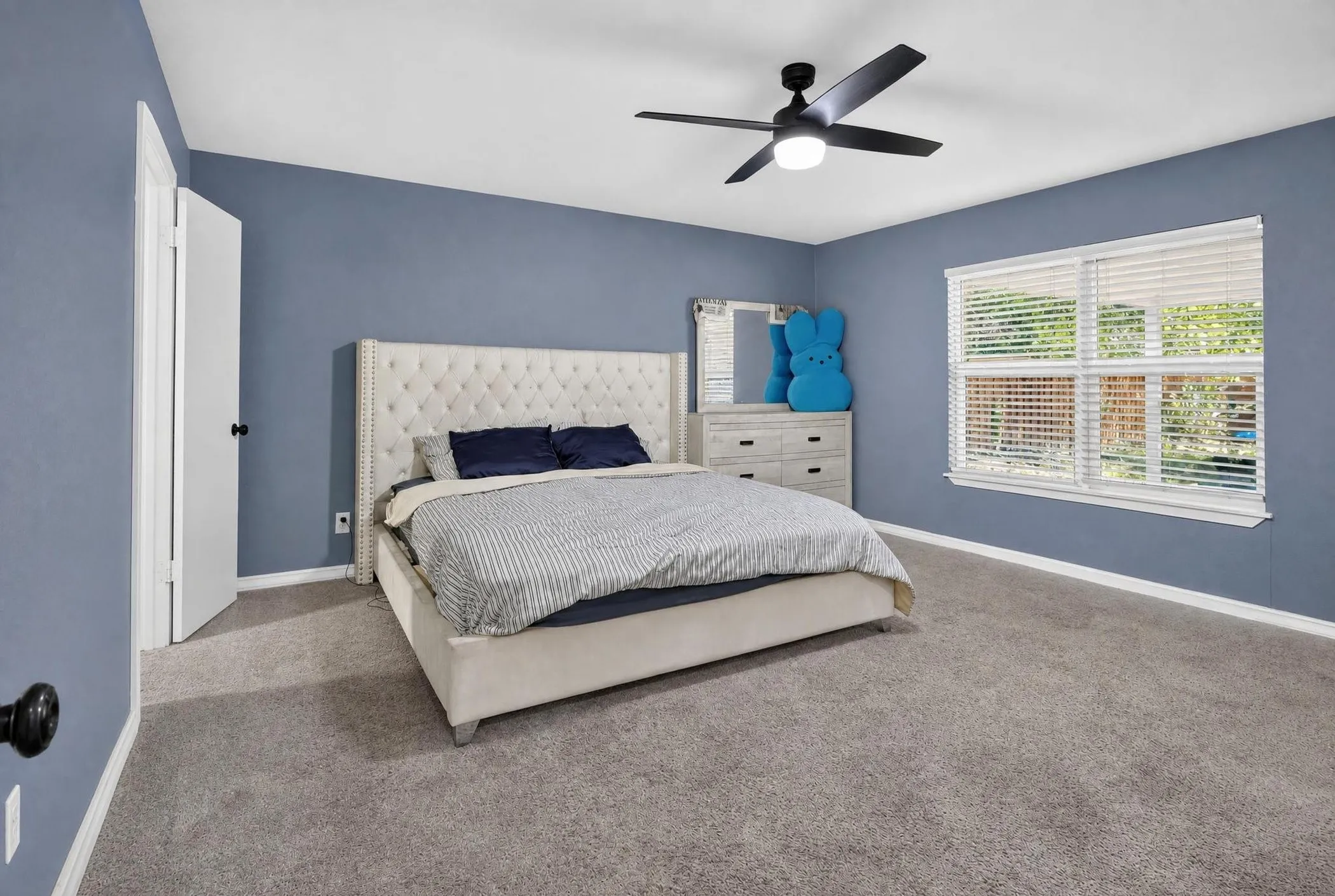 Spacious primary bedroom with soft blue walls, carpet flooring, and abundant natural light from dual windows with plantation shutters