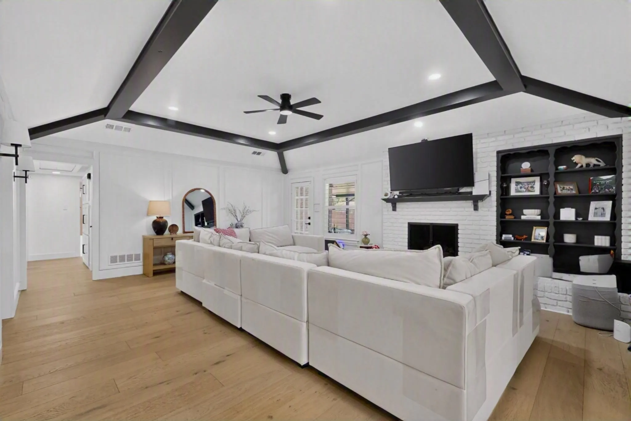 Wood-finish flooring, exposed ceiling beams, built-in shelving, white brick fireplace, and paneling detailing