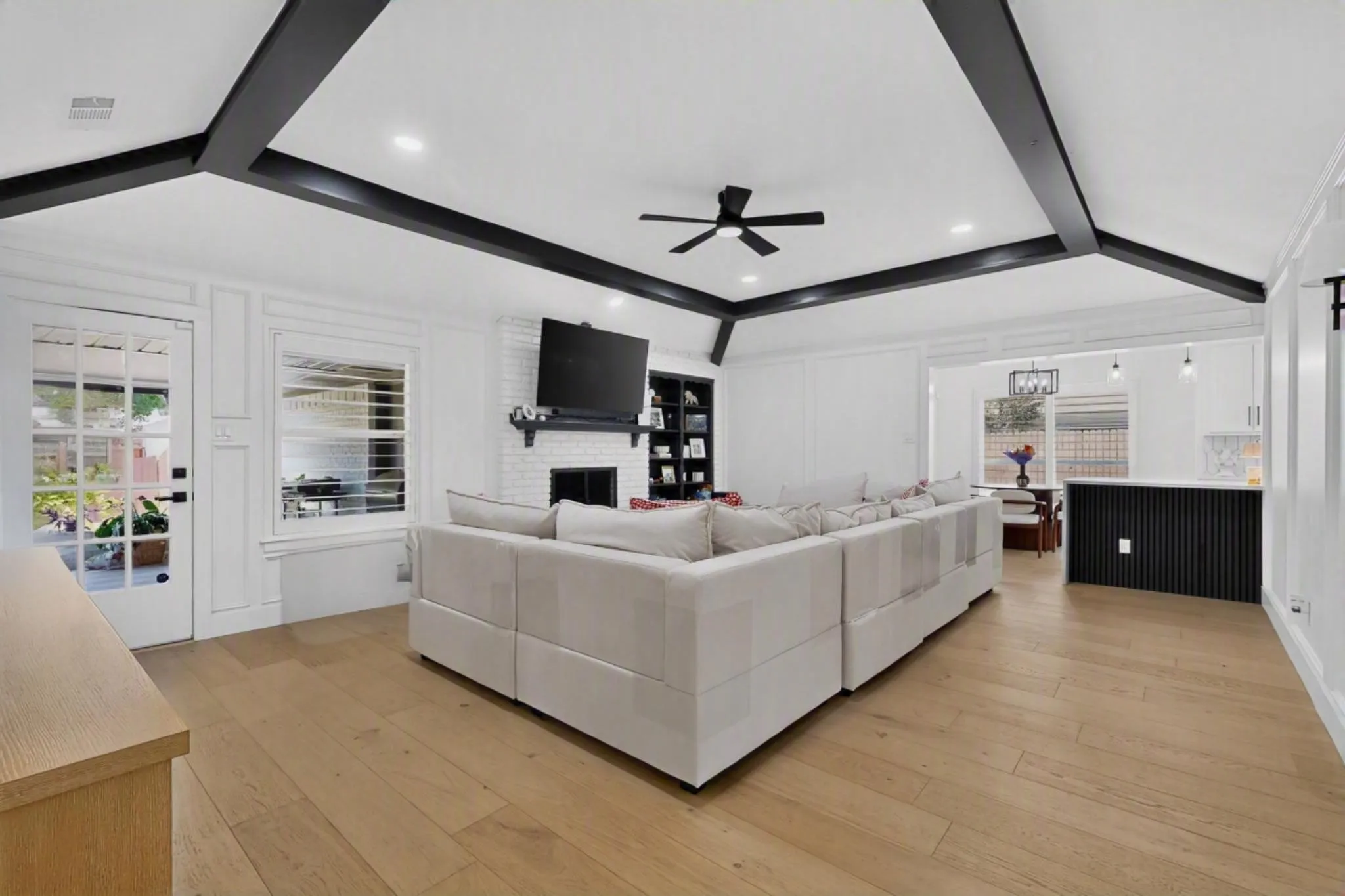 Vaulted ceiling with exposed black beams, light wood-finish flooring, white brick gas fireplace, built-in shelving, and a black fluted kitchen island