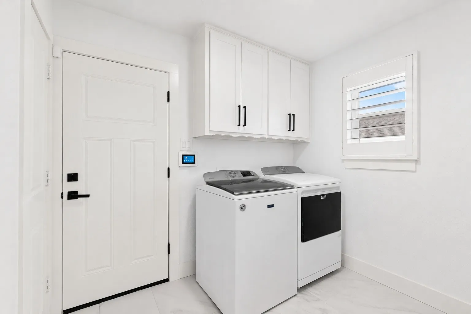 White paneled door with matte black hardware, white upper cabinetry with matte black pulls, and a window with white plantation shutters