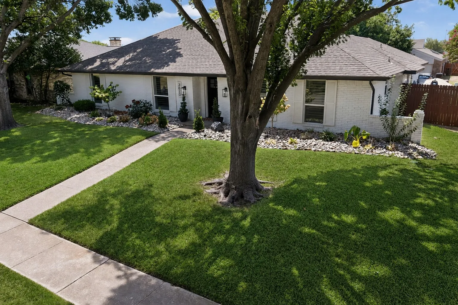 White brick exterior, dark composition shingle roof, well-defined landscaping beds, mature trees, and a concrete walkway