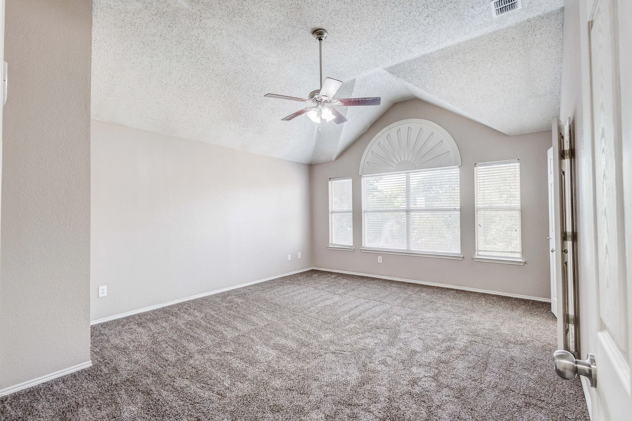 Spacious room featuring vaulted ceilings, a prominent arched window, two additional rectangular windows with blinds, plush gray carpet, and a ceiling fan