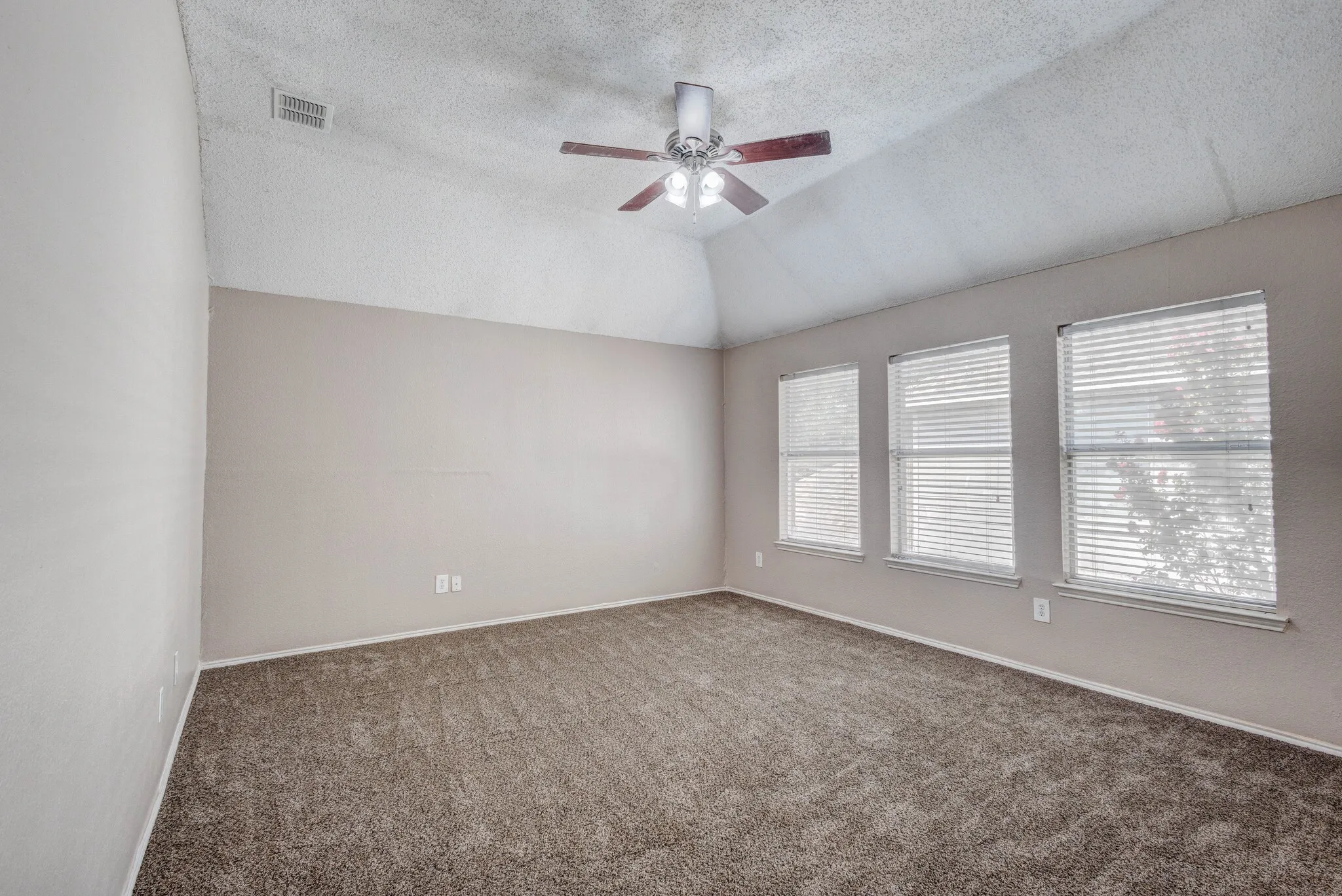 Carpeted interior space featuring a vaulted ceiling, three windows with blinds, a ceiling fan with integrated lighting, and neutral wall tones