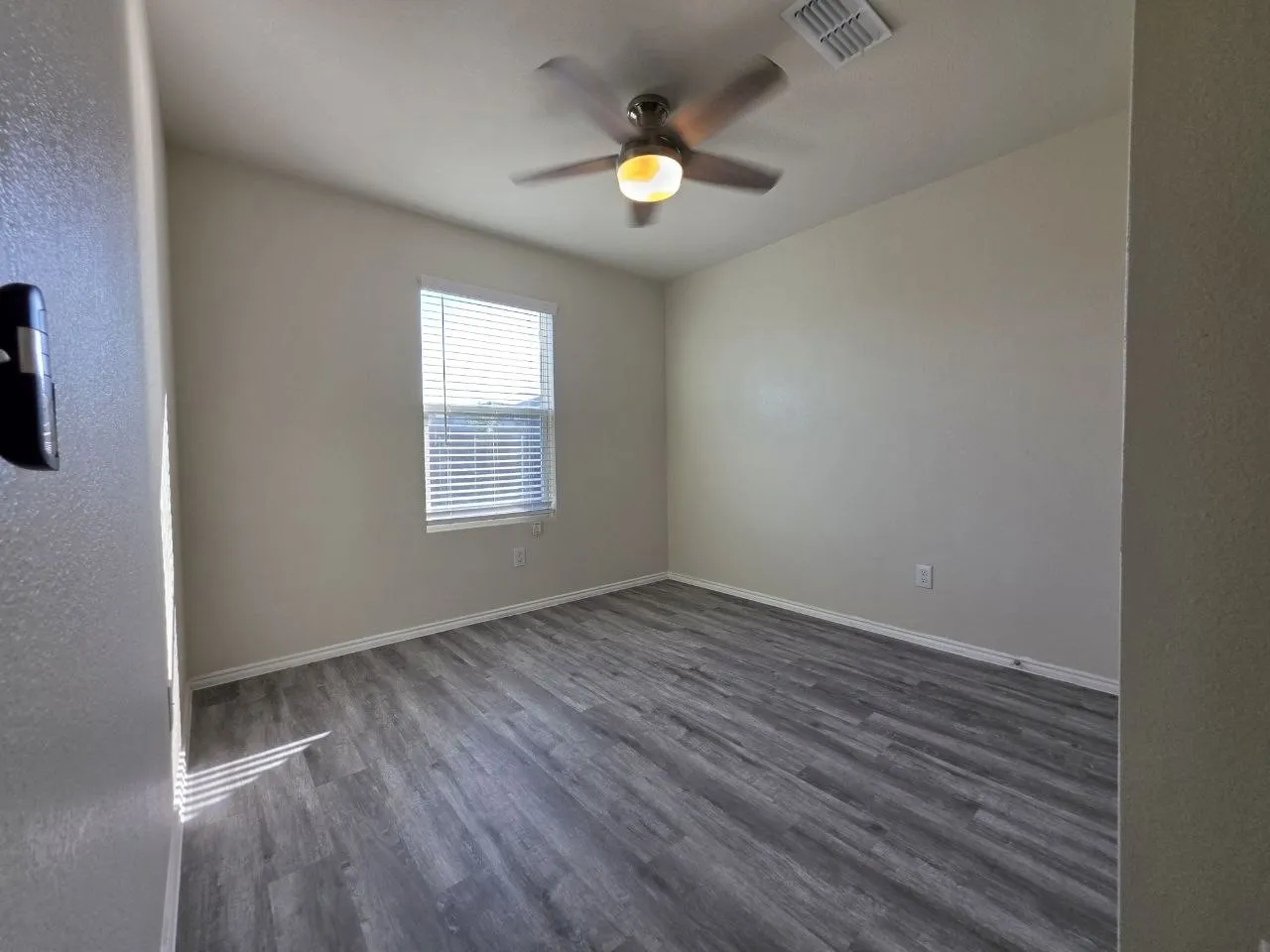 Gray wood-finish flooring, neutral wall tones, window with horizontal blinds, and a ceiling fan with integrated lighting