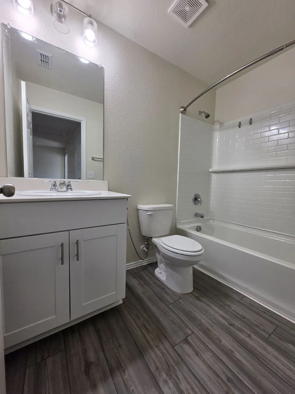 Wood-finish flooring, white vanity with dual doors, subway tile shower surround, chrome plumbing fixtures, and wall-mounted light fixture