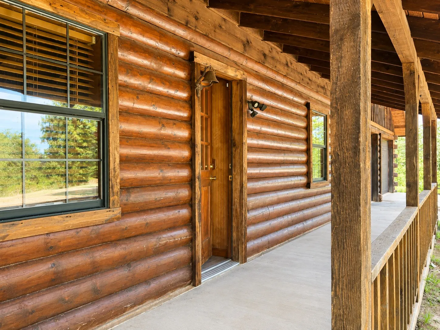 Expansive covered porch featuring a log cabin exterior, exposed wood beam ceiling, concrete flooring, black-framed windows, and a solid wood entry door