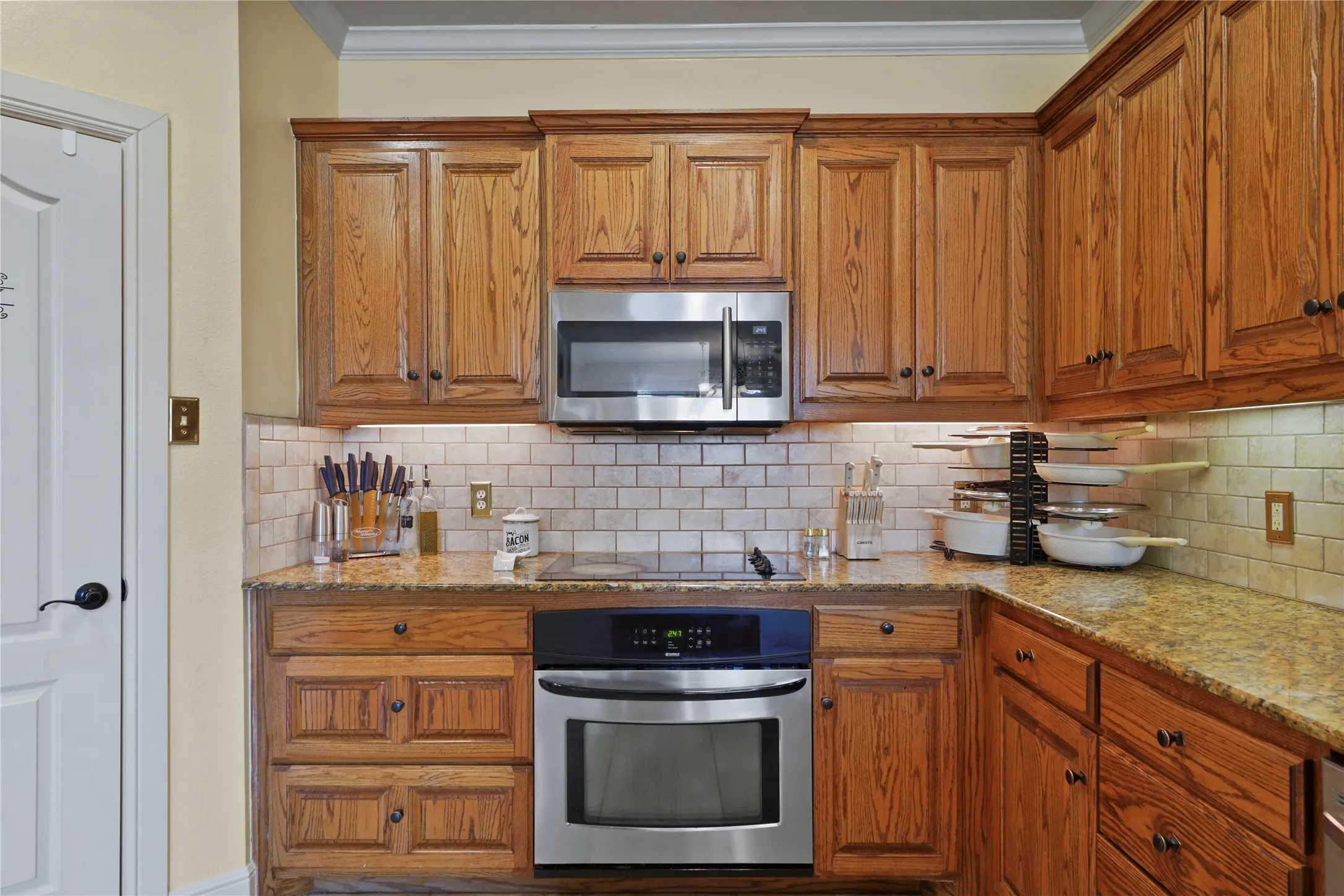 Wood-finish cabinetry with crown molding, stainless steel microwave, stainless steel wall oven, light-colored subway tile backsplash, and stone countertops