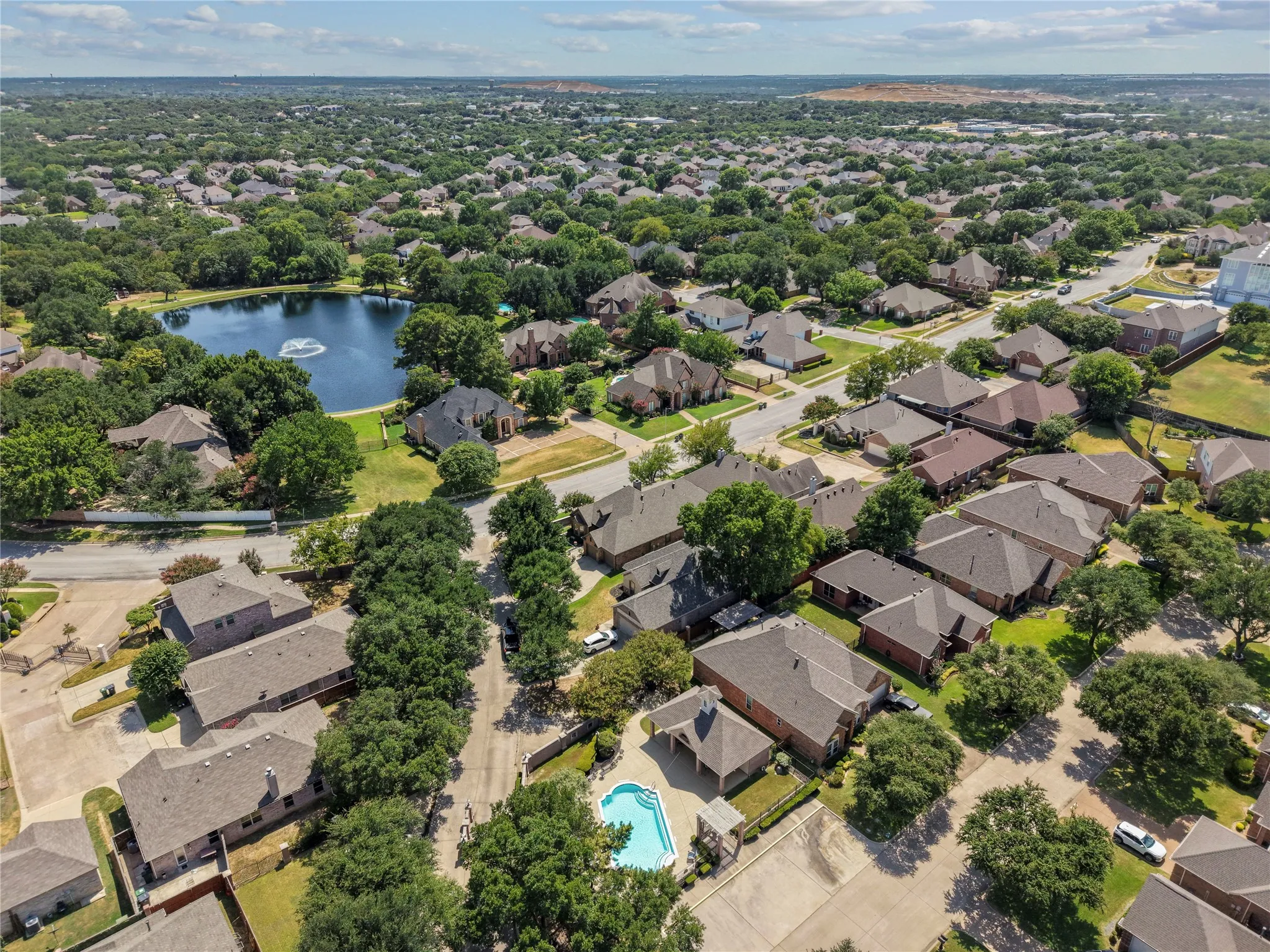 Expansive residential area featuring numerous single-family homes, mature tree coverage, a prominent central pond with a fountain, and several private swimming pools