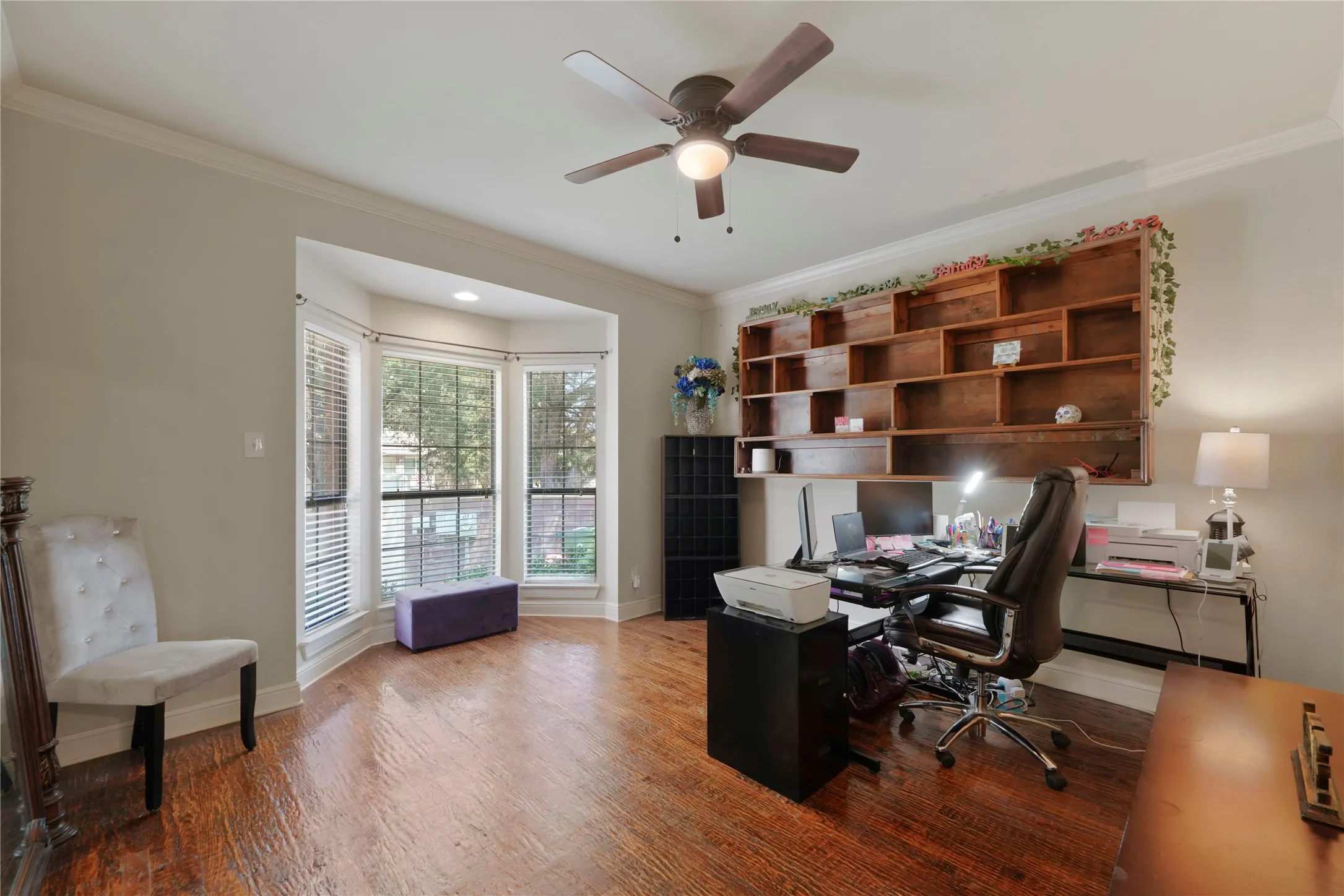 Bay window with blinds, wood-finish flooring, built-in shelving, ceiling fan with integrated lighting, and crown molding